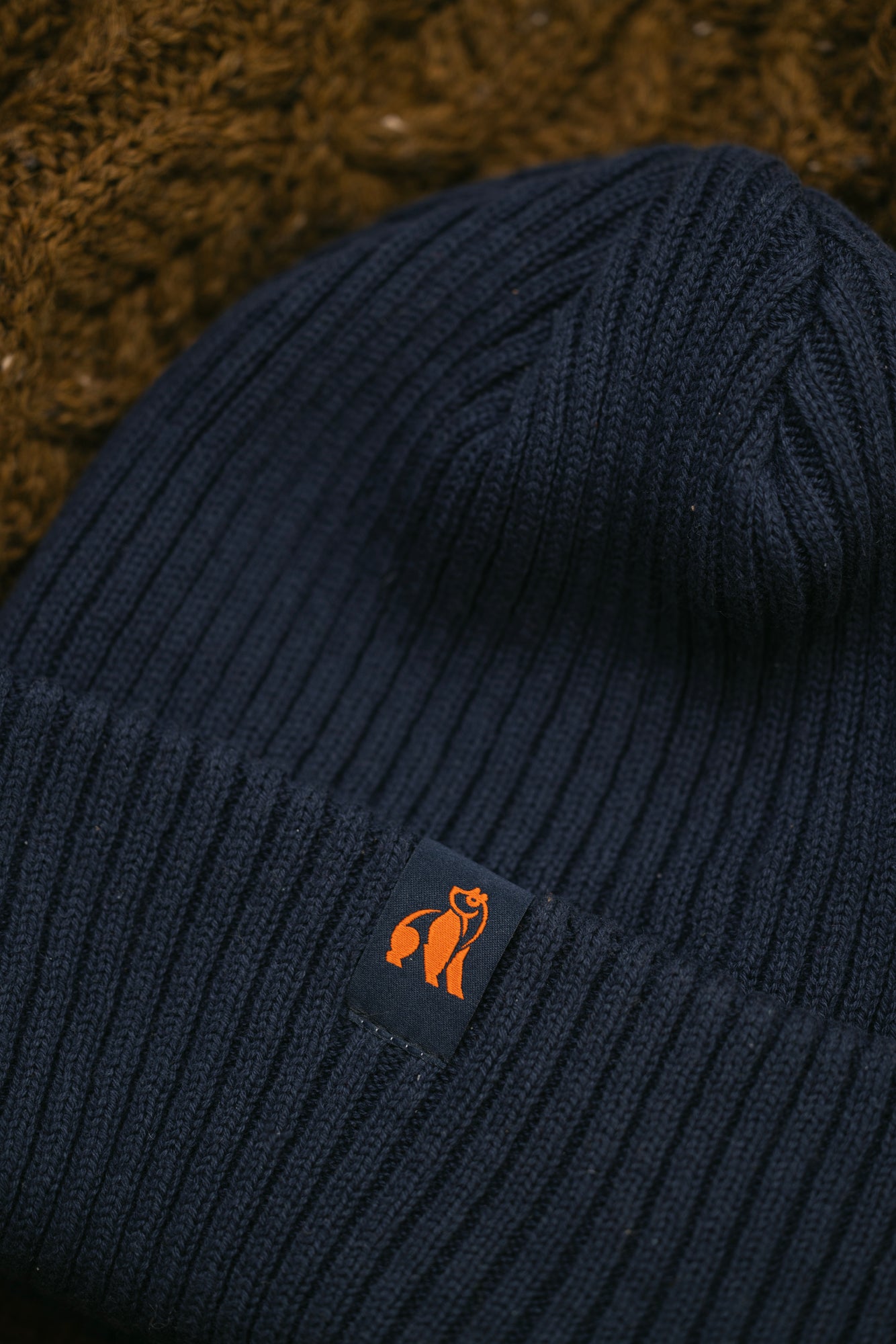 Close up of a dark navy blue ribbed knit beanie with an orange Swole Panda logo tag.