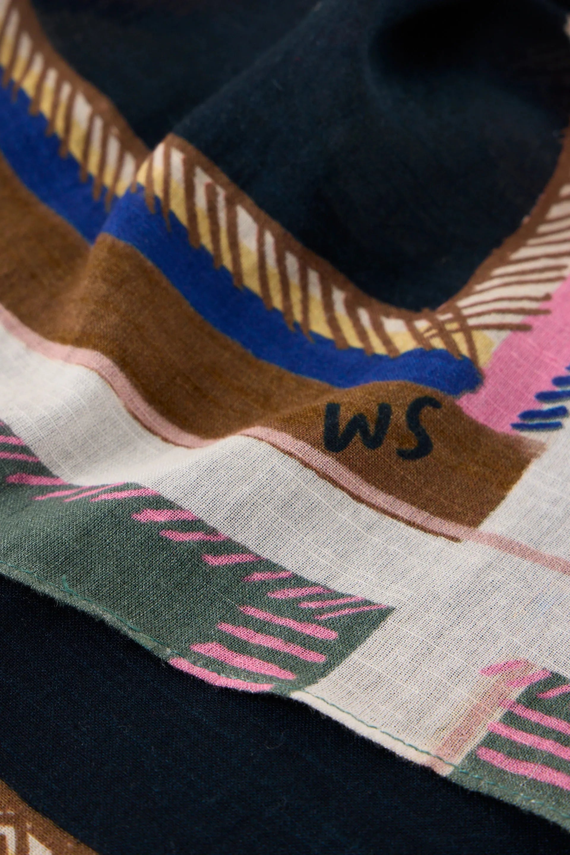 A close-up of a patterned scarf featuring stripes and geometric shapes in brown, pink, blue, green, and cream, with the letters 'WS' embroidered on a brown section.