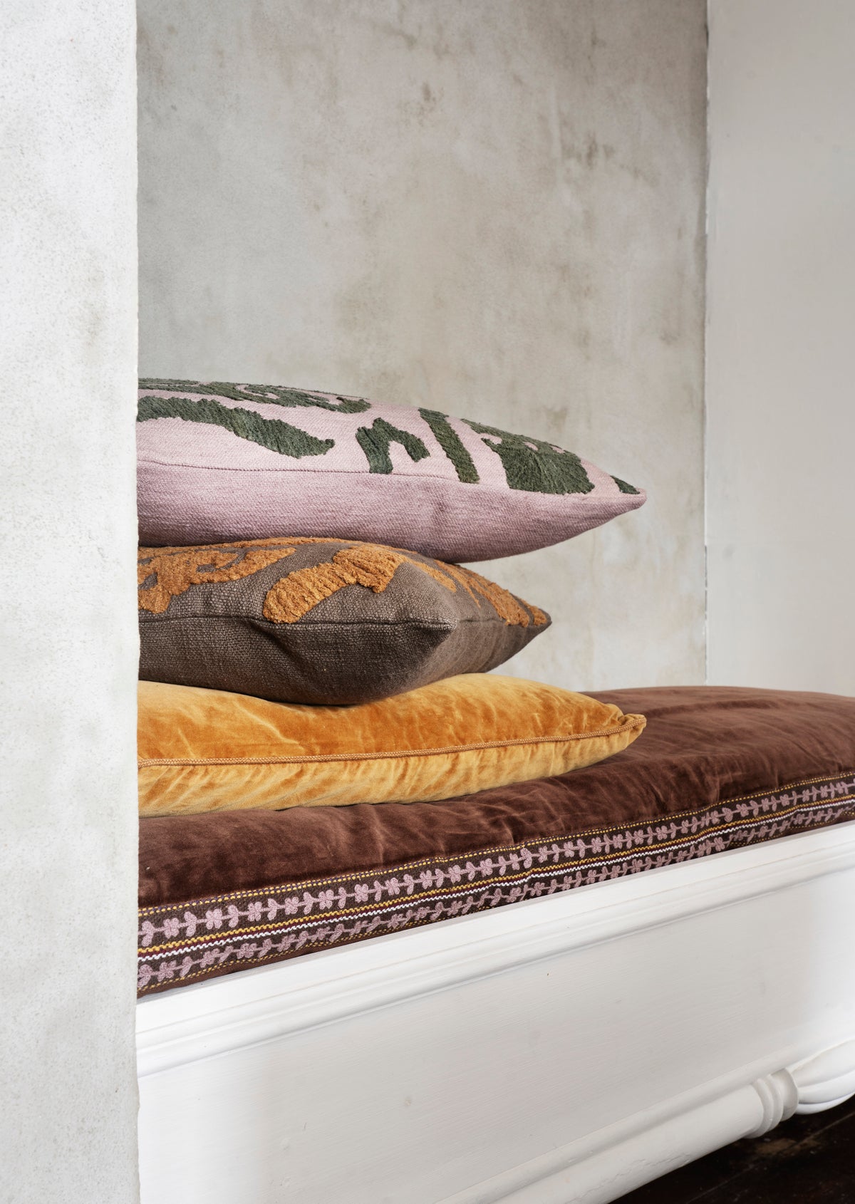 Three decorative throw pillows stacked on a brown upholstered bench in a room with a textured light gray wall.