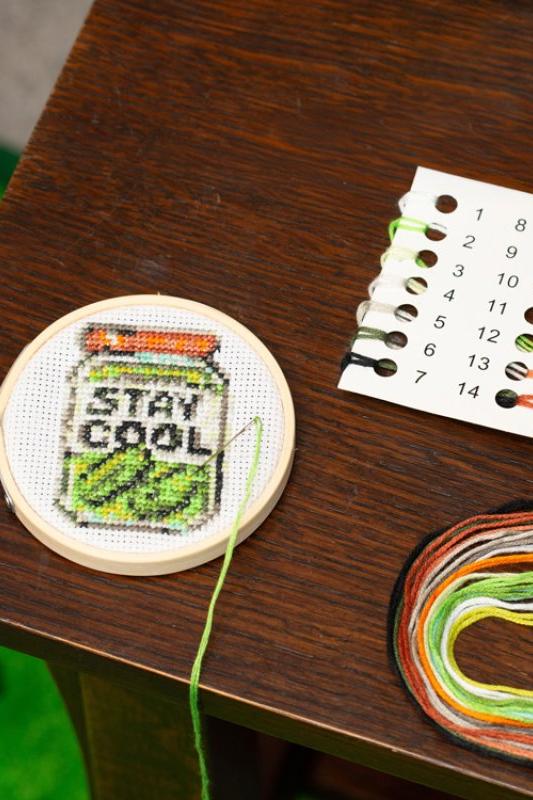 Embroidery hoop with 'Stay Cool' text and a jar design on a wooden table, with embroidery threads and a color chart.