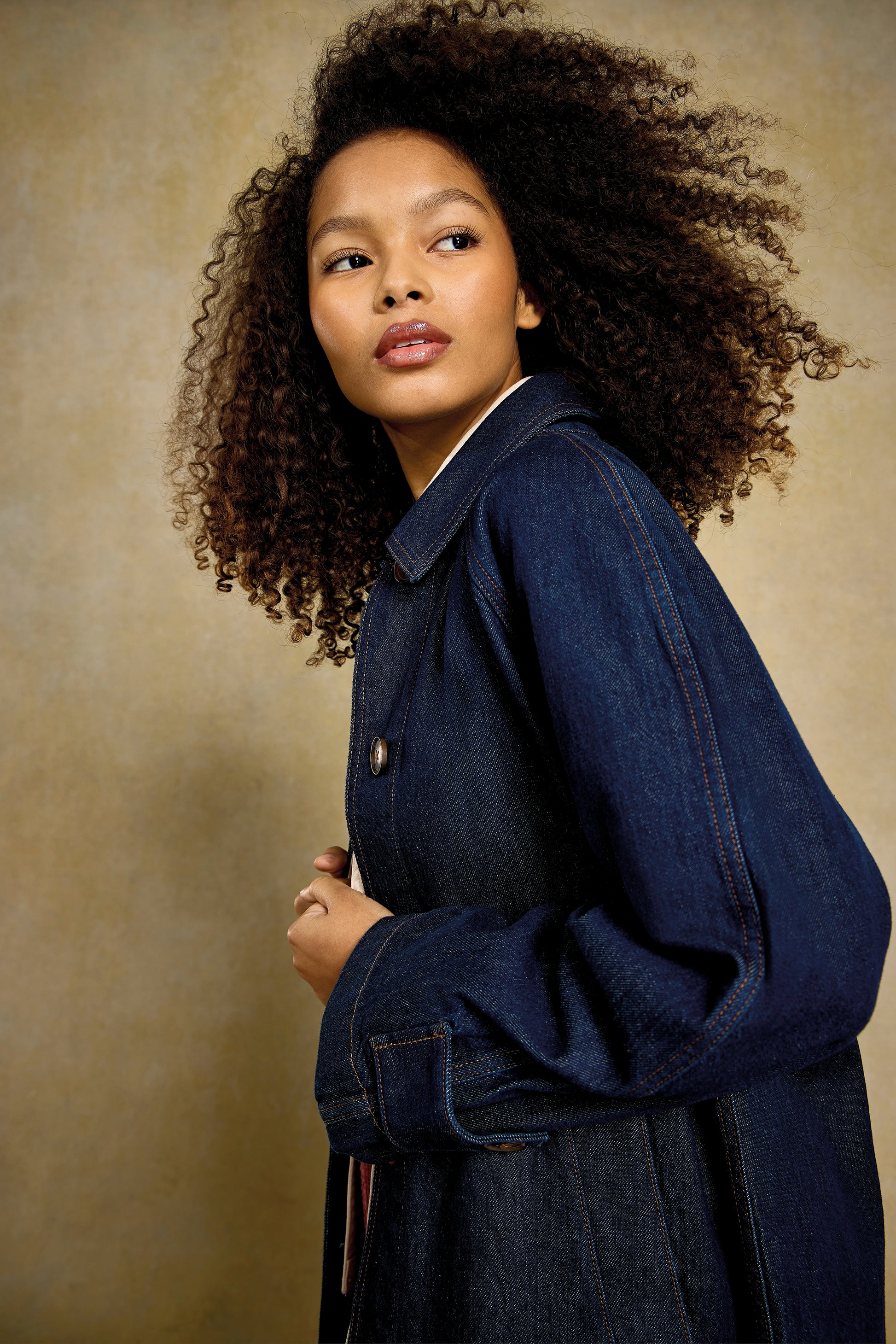 Woman with curly hair wearing a dark denim jacket looking over her shoulder
