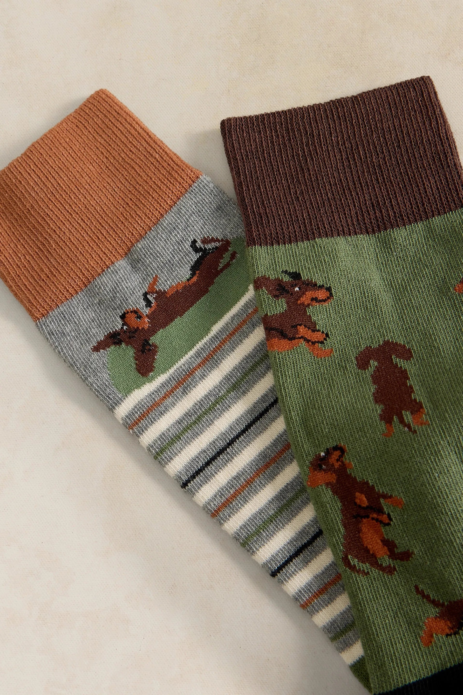 Two pairs of patterned crew socks laid flat on a light beige surface. One pair has a brown cuff and a green body with brown dog illustrations, plus white and gray stripes near the foot.