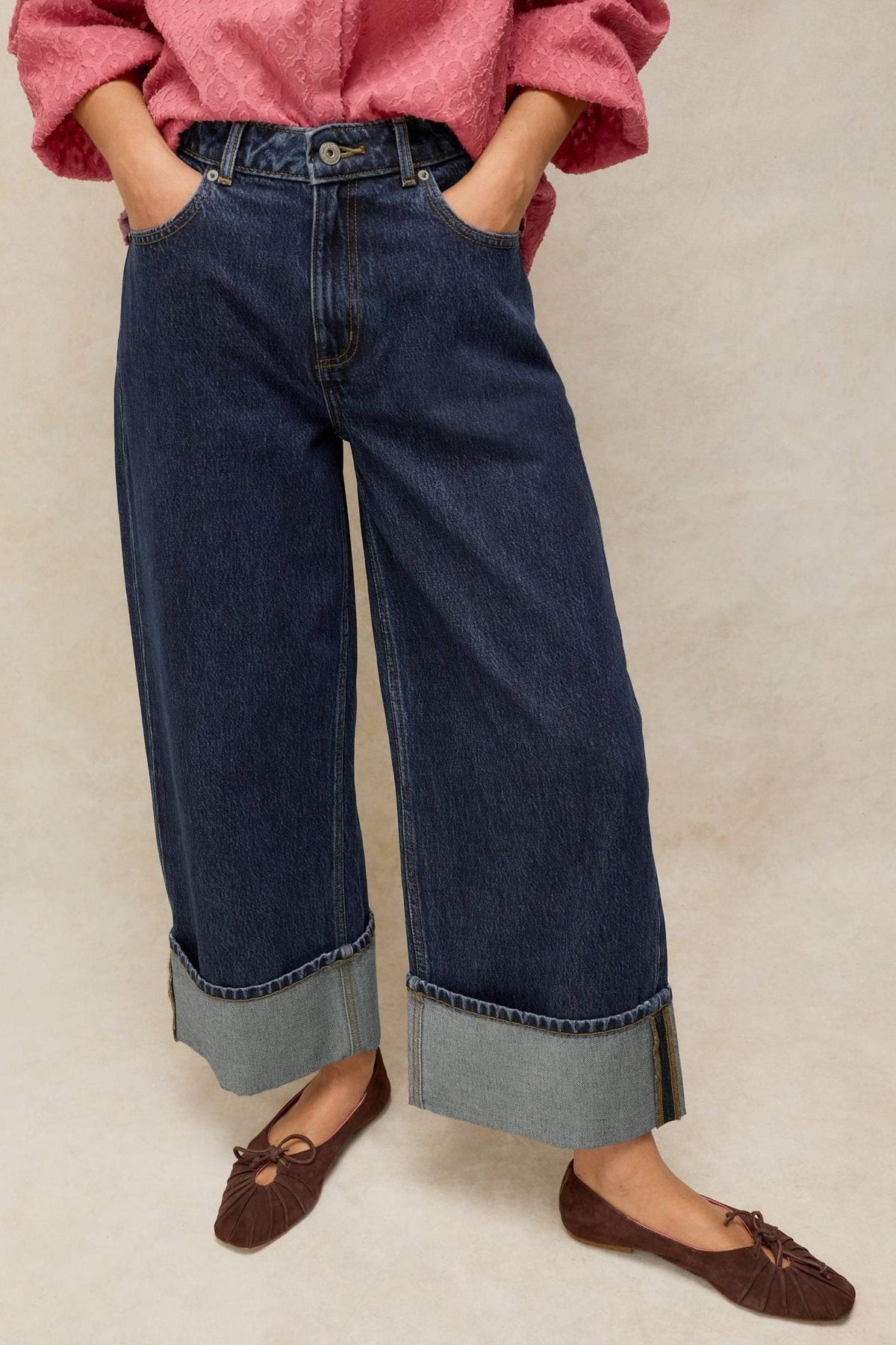 A person wearing dark denim jeans with wide cuffed hems, a pink textured top, and brown ballet flats.