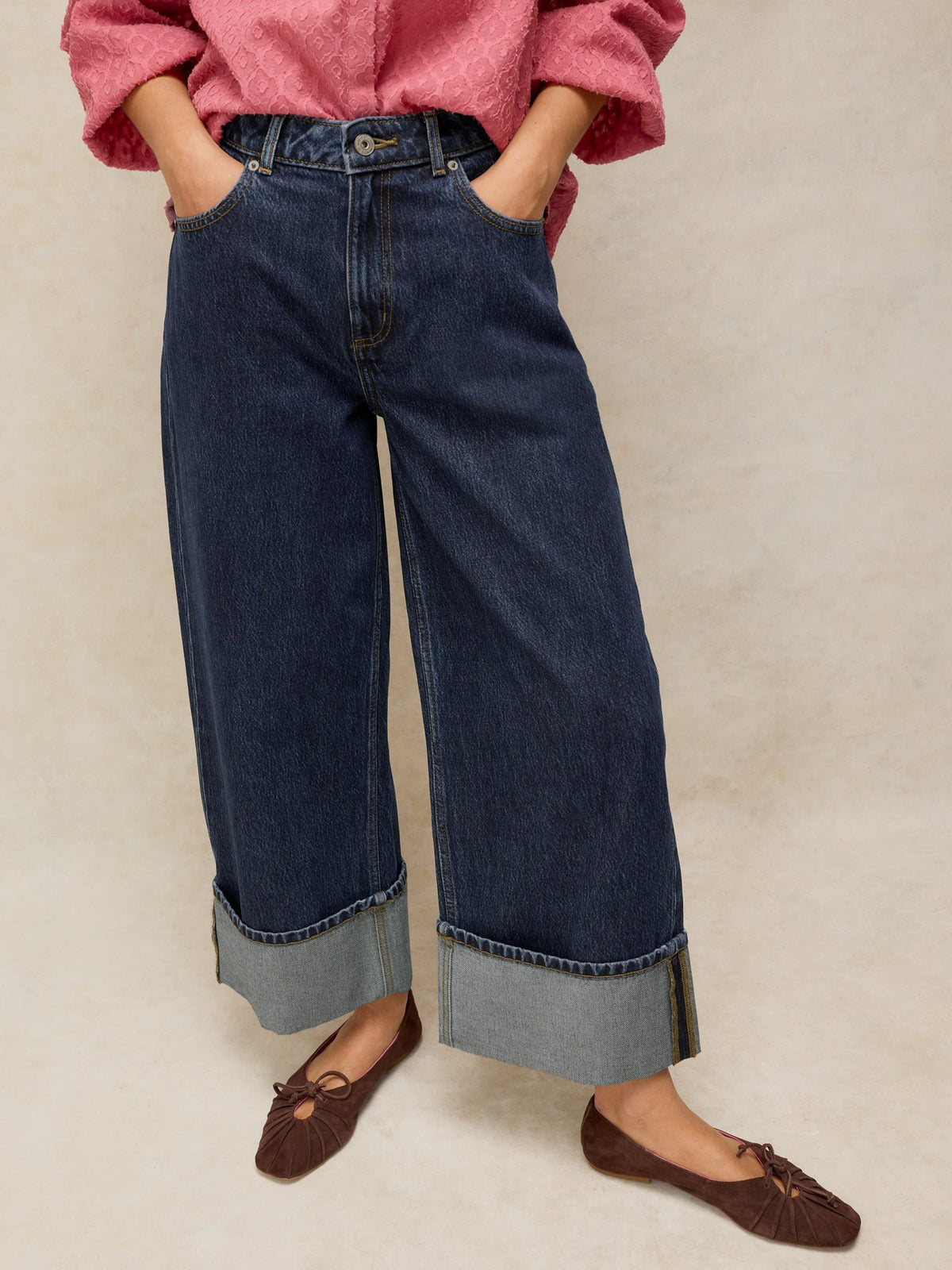 A person wearing dark denim jeans with wide cuffed hems, a pink textured top, and brown ballet flats.
