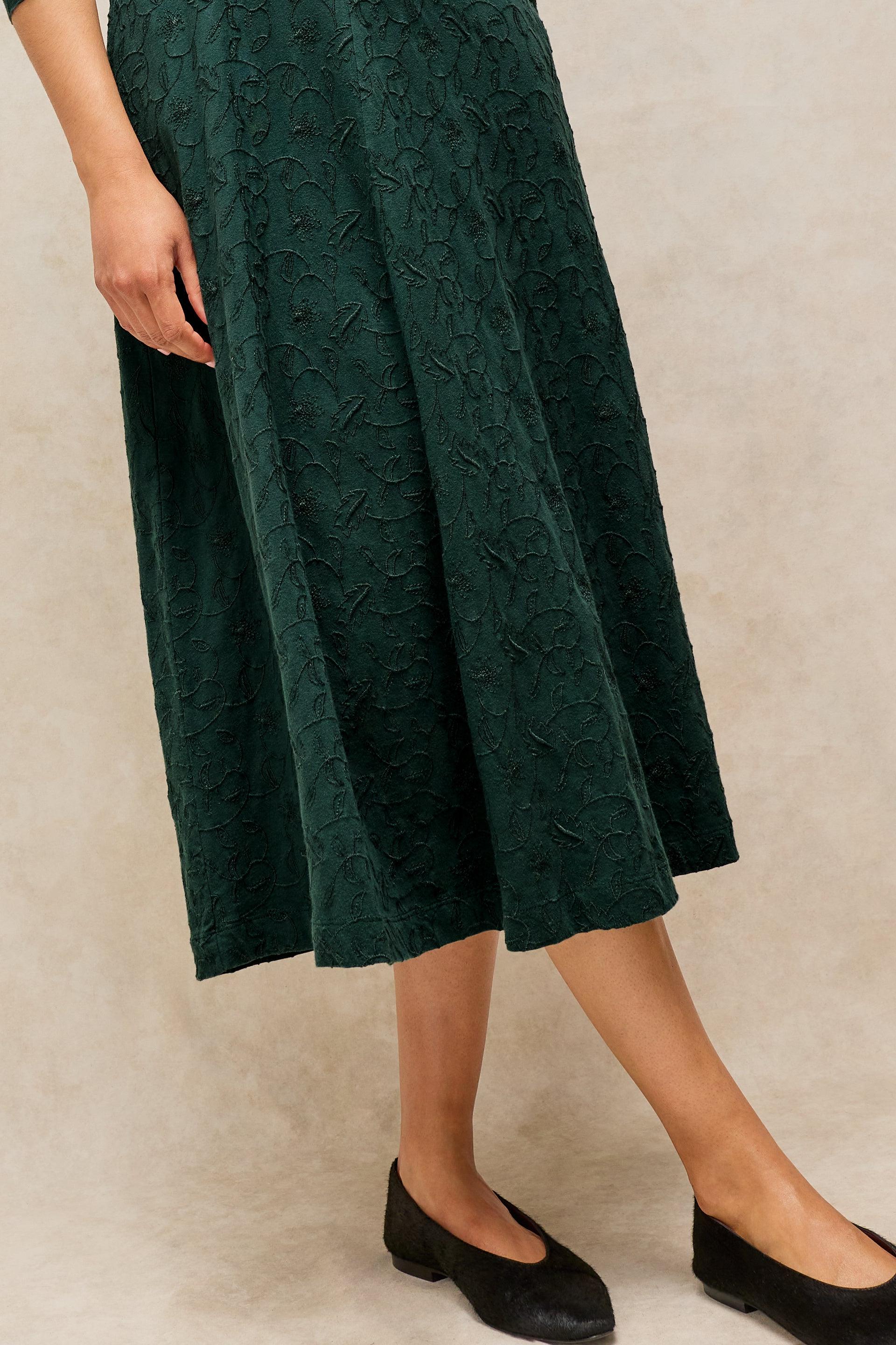 A dark green midi skirt with a textured floral jacquard pattern, shown on a model wearing black flats against a neutral beige background.