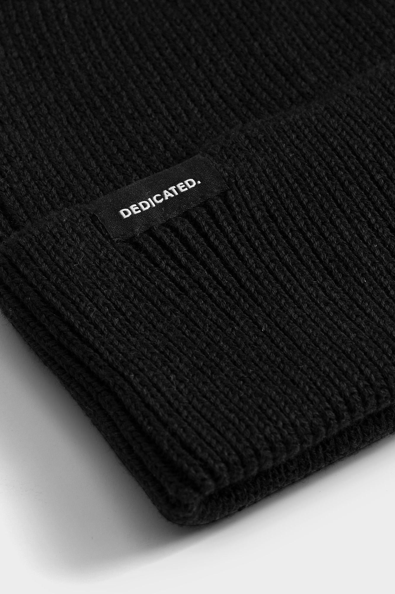 Close-up of a black ribbed knit beanie with a folded cuff and a small black label reading DEDICATED. in white text, photographed on a light gray background.