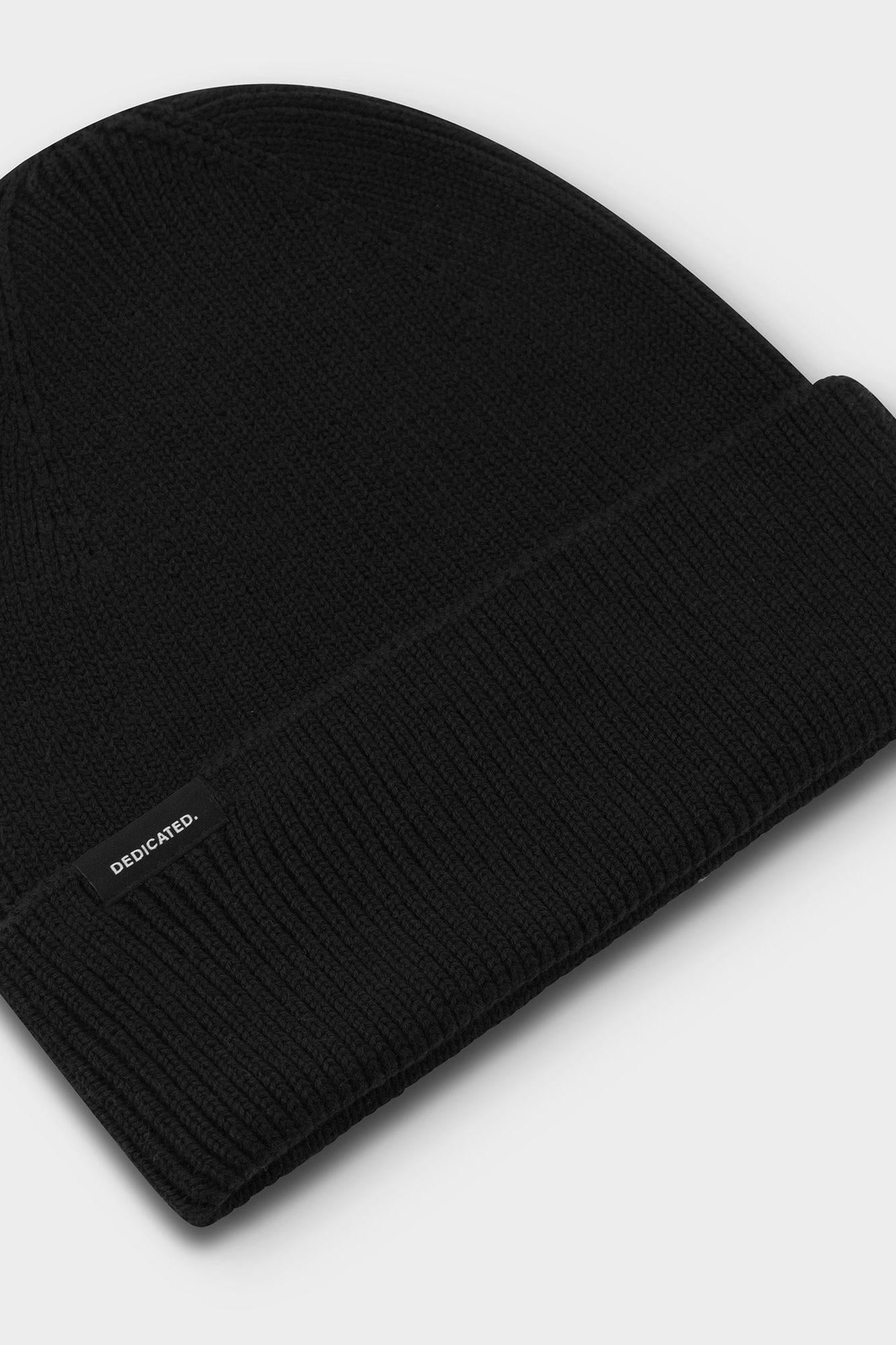 A black ribbed knit beanie hat with a folded cuff, featuring a small white 'DEDICATED.' brand tag on the side, set against a light gray background.