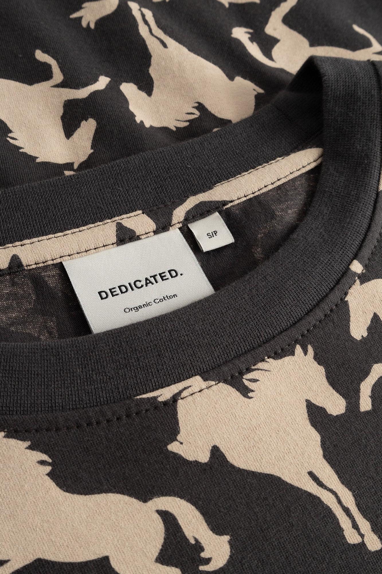 Close-up of a DEDICATED. organic cotton t-shirt featuring a dark charcoal gray background with a beige silhouette print of running horses, showing the inner collar and size tag.