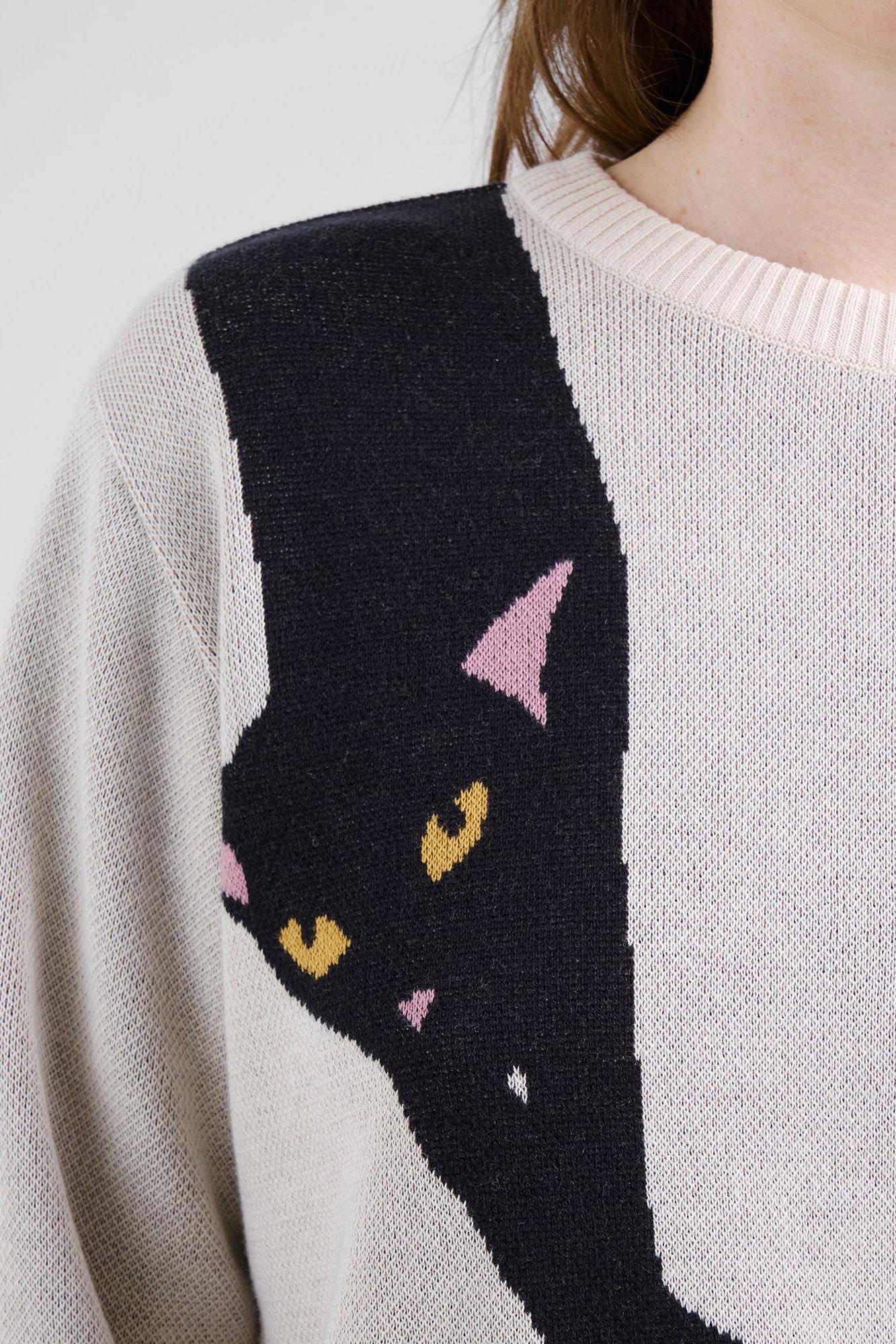 Close-up of a person wearing a knit sweater with a large black cat graphic across the chest and shoulder, with pink ears and yellow eyes, on a light gray background.
