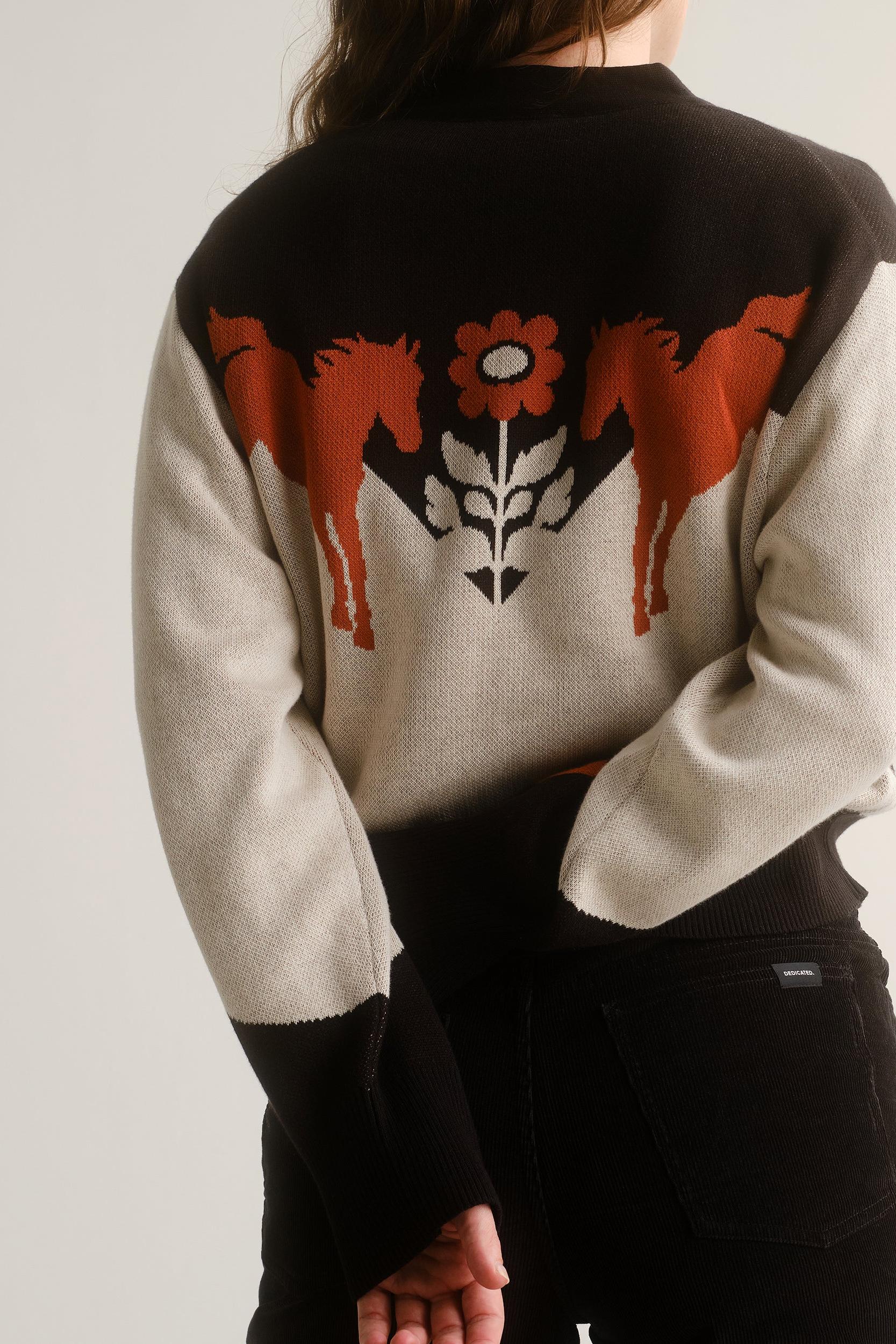 A person shown from the back wearing a knit sweater with a large graphic design. The sweater has a black upper yoke and black ribbed collar/cuffs, off-white sleeves and lower body, and a central motif featuring orange horses, a red flower, and a white wolf head.