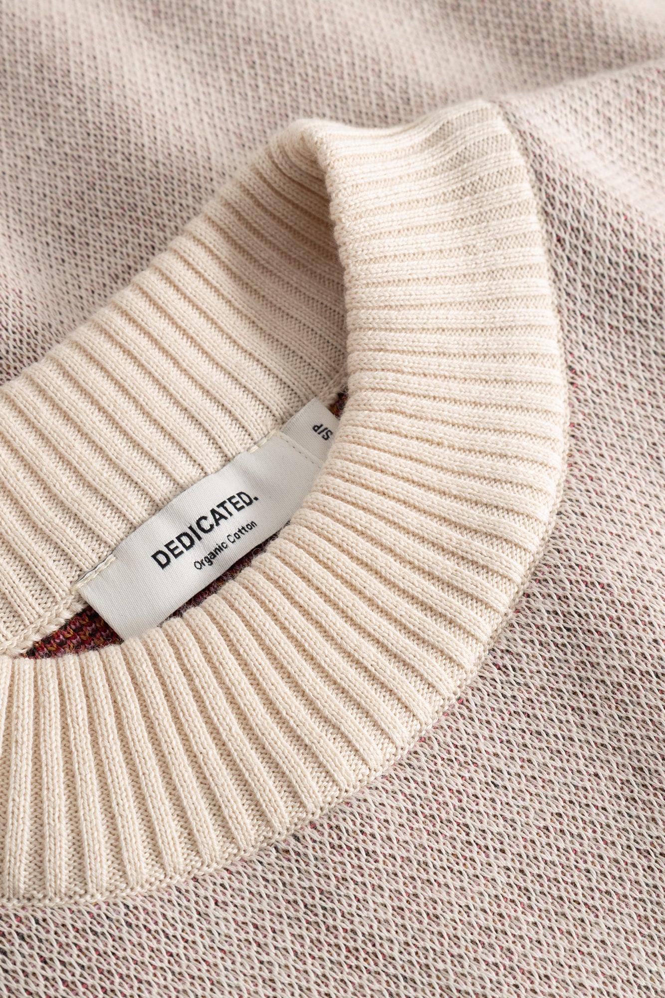 Close-up of a cream-colored knit crewneck sweater showing the ribbed collar and a white brand label that reads 'DEDICATED Organic Cotton'.
