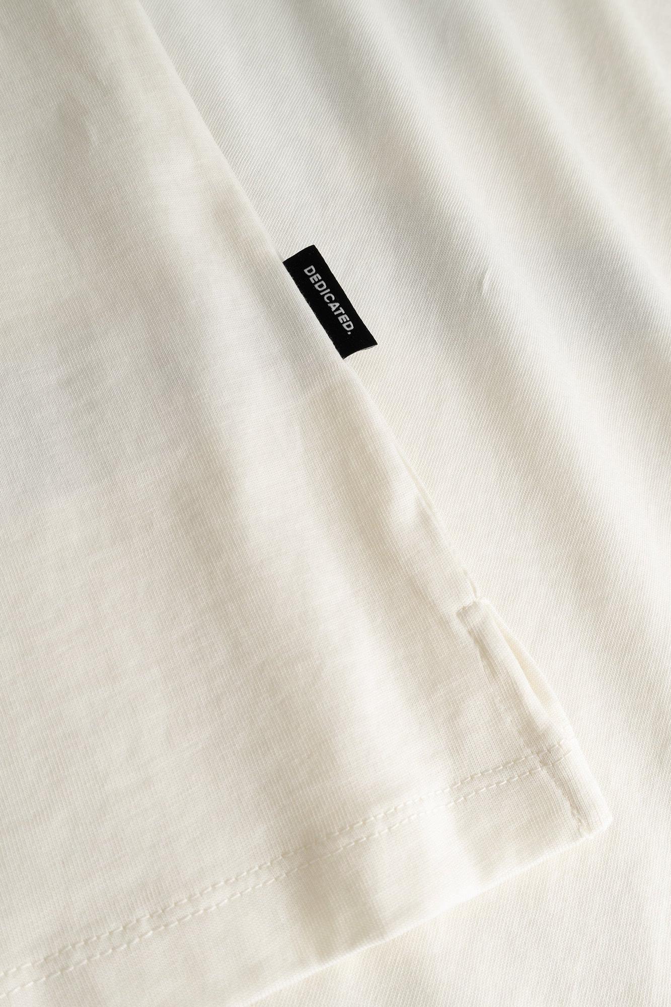 Close-up of an off-white cotton t-shirt fabric with a small black rectangular brand tag reading 'Dedicated.' sewn near the shoulder seam.
