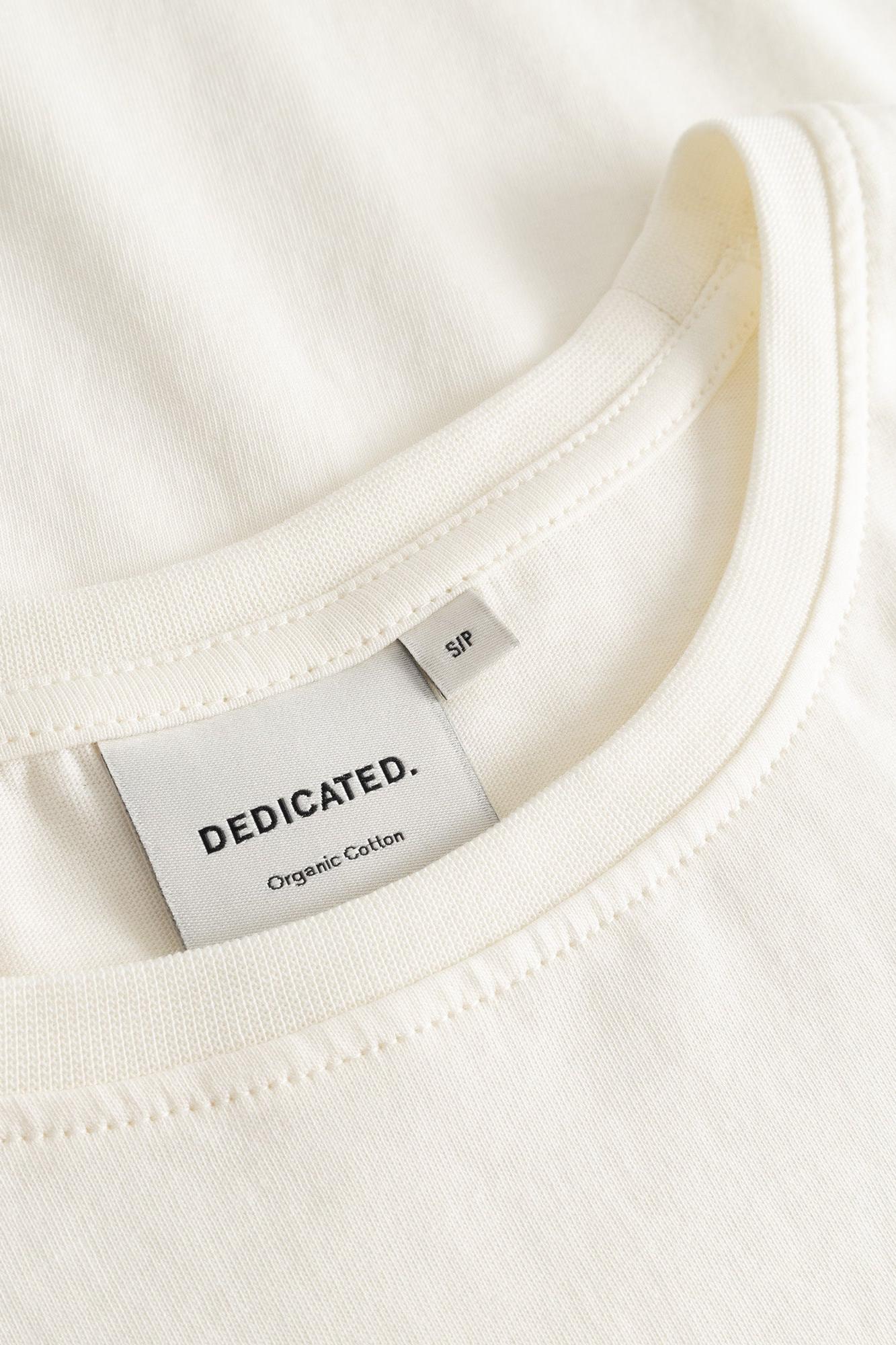 Close-up of a cream-colored crewneck t-shirt showing the inner collar with a white 'DEDICATED. Organic Cotton' brand label and a small size tag reading 'SFP'.