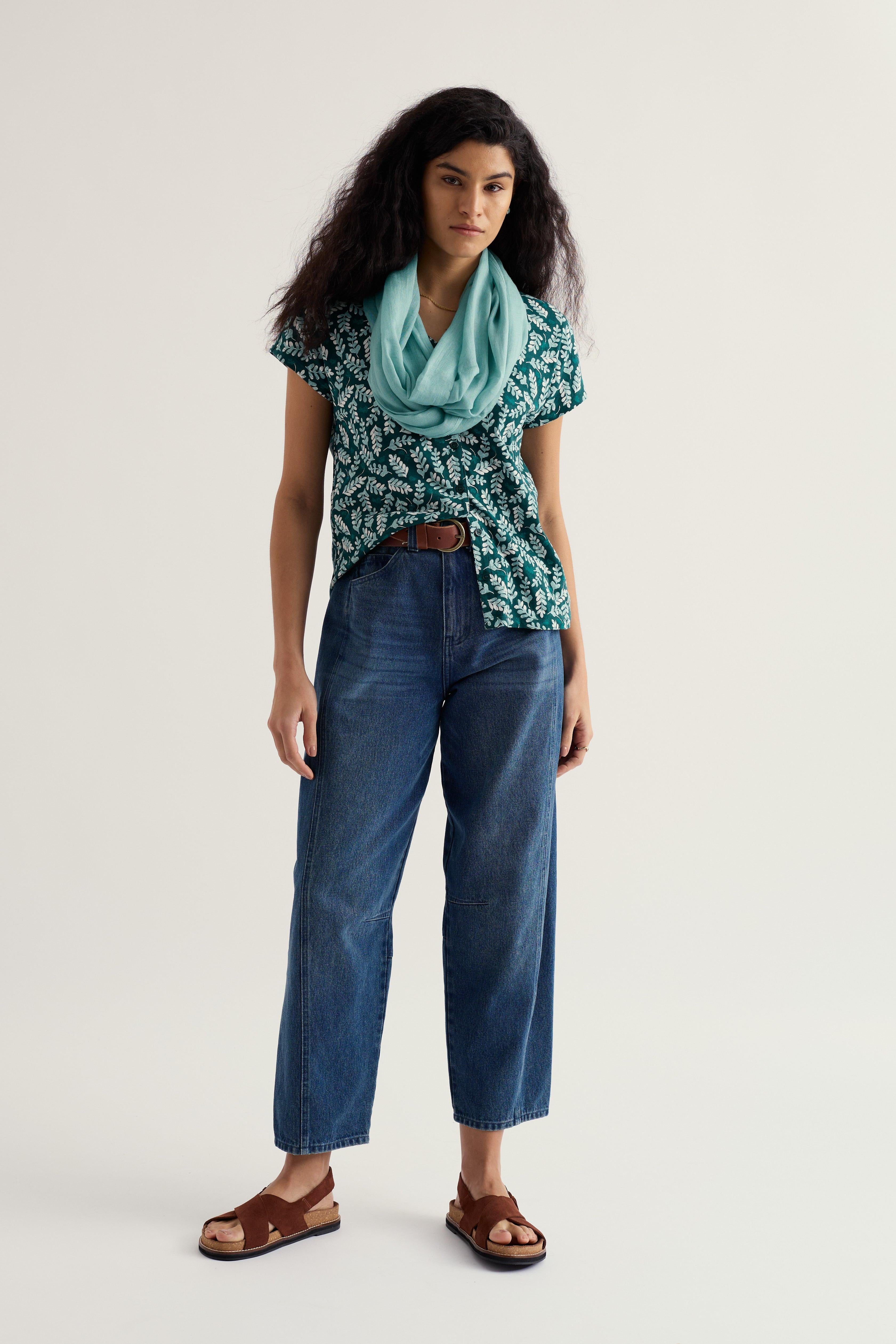 Woman wearing a patterned organic cotton top, blue jeans, and a light blue scarf on a plain background