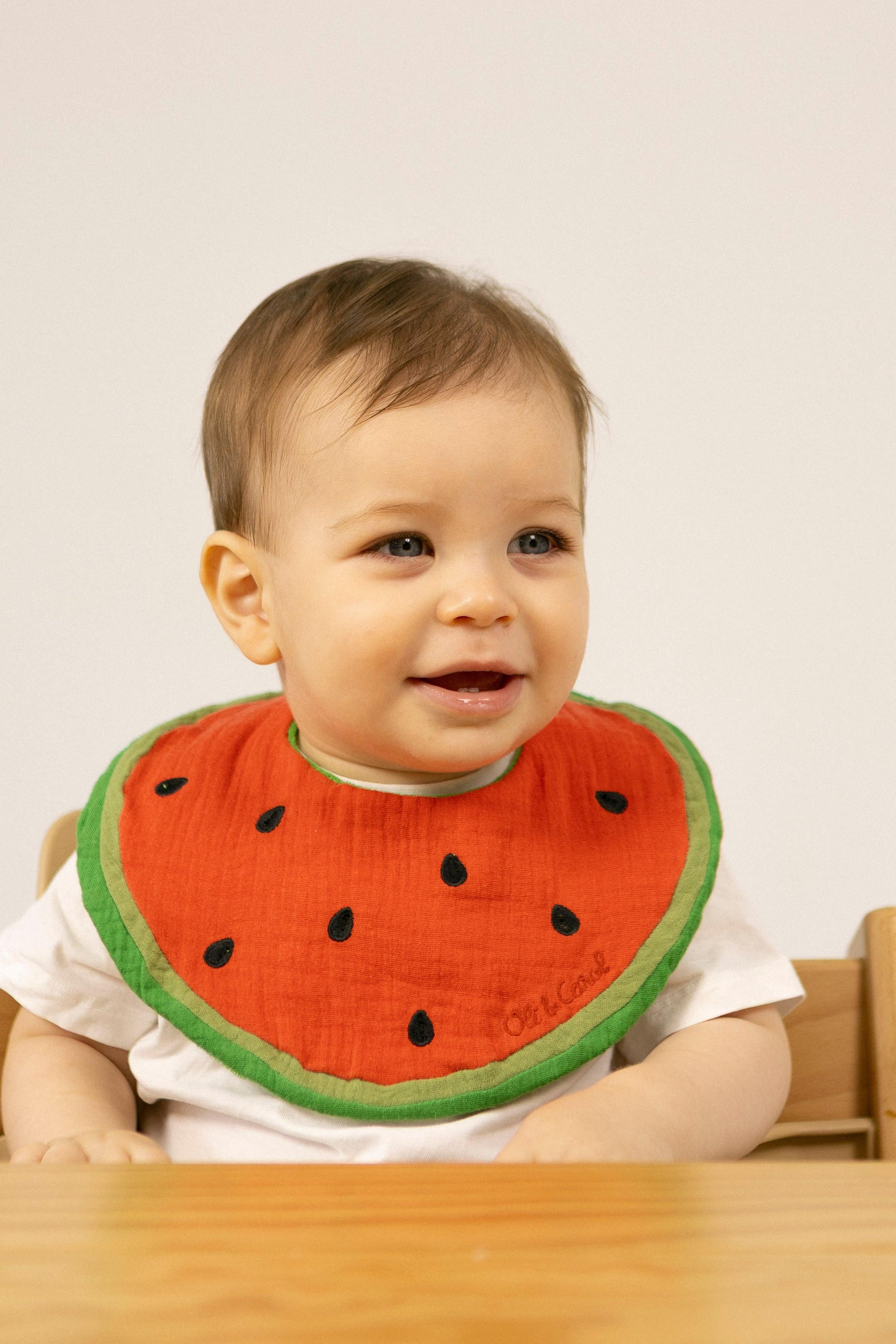 Baby wearing an organic cotton baby bib with fun watermelon design with black seeds on a plain background
