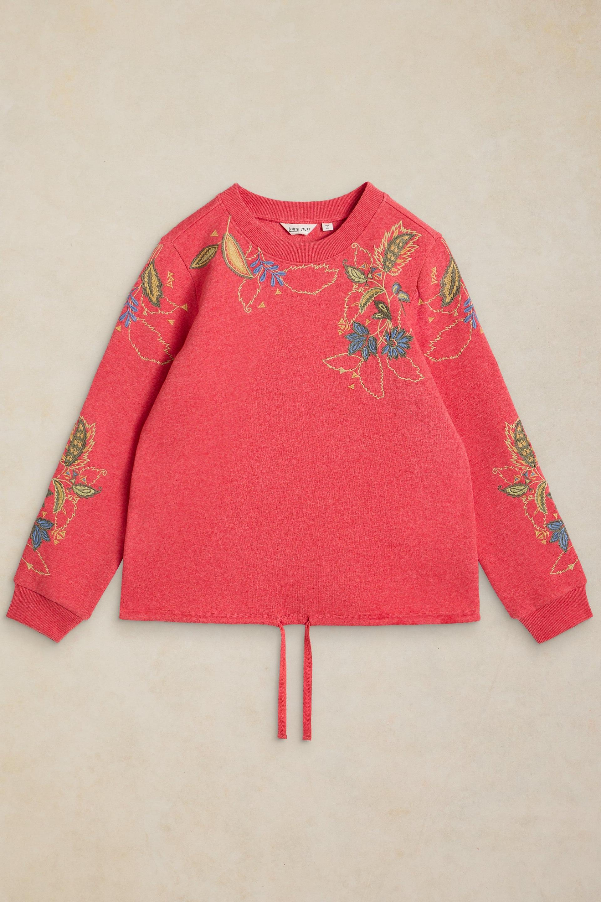 A coral pink long-sleeve knit sweater with colorful floral embroidery on the chest and sleeves, featuring a drawstring hem, laid flat on a textured beige background.