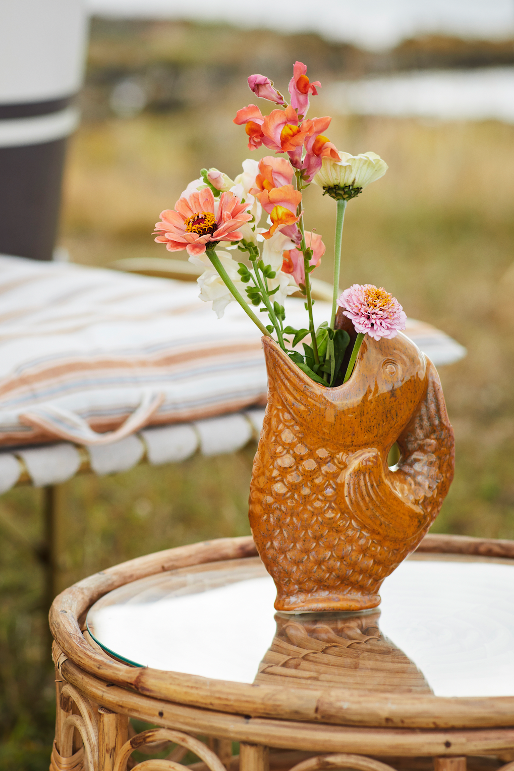 A ceramic fish-shaped vase filled with colorful snapdragons and zinnias on a glass-topped rattan table.