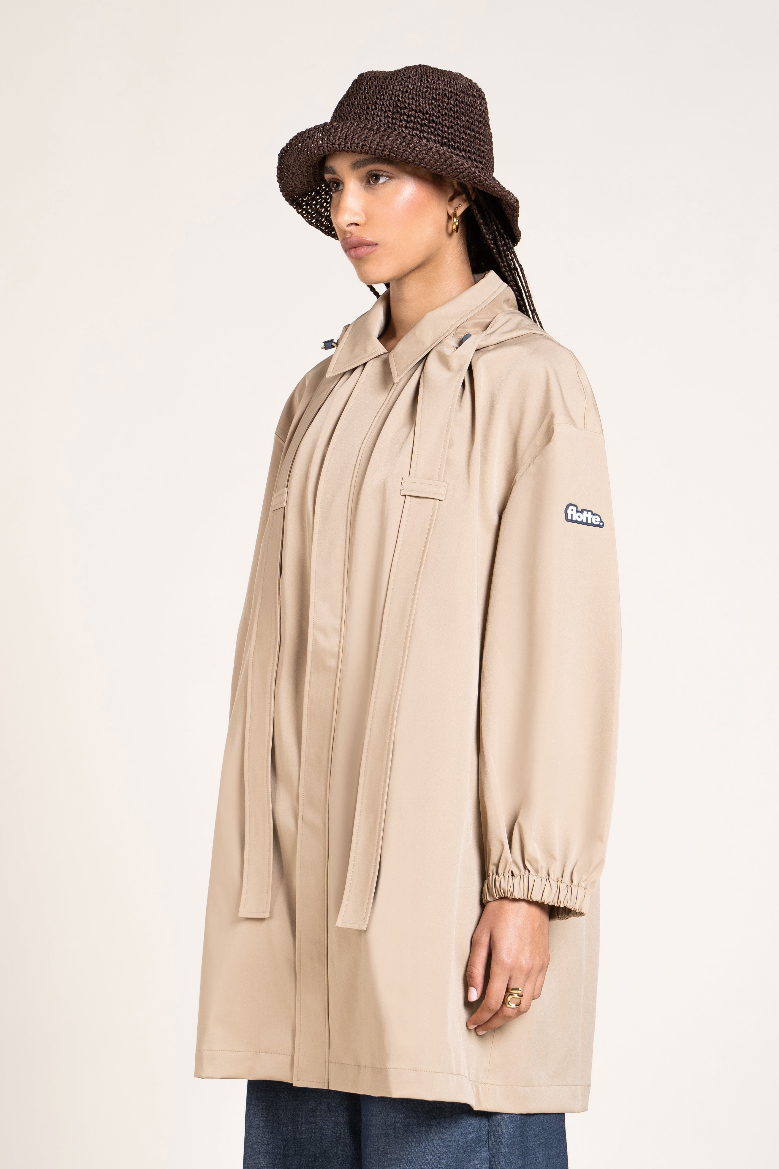 A model wearing a beige Flotte waterproof coat and a brown crochet bucket hat against a plain background.