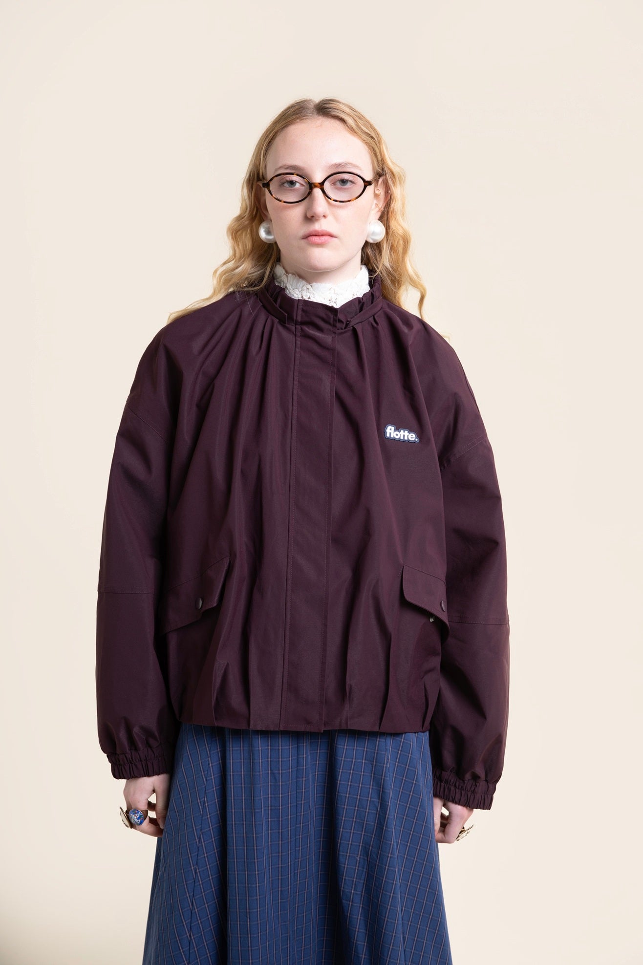 A blonde model wearing a dark purple Flotte waterproof coat over a white lace collar and a blue plaid skirt.