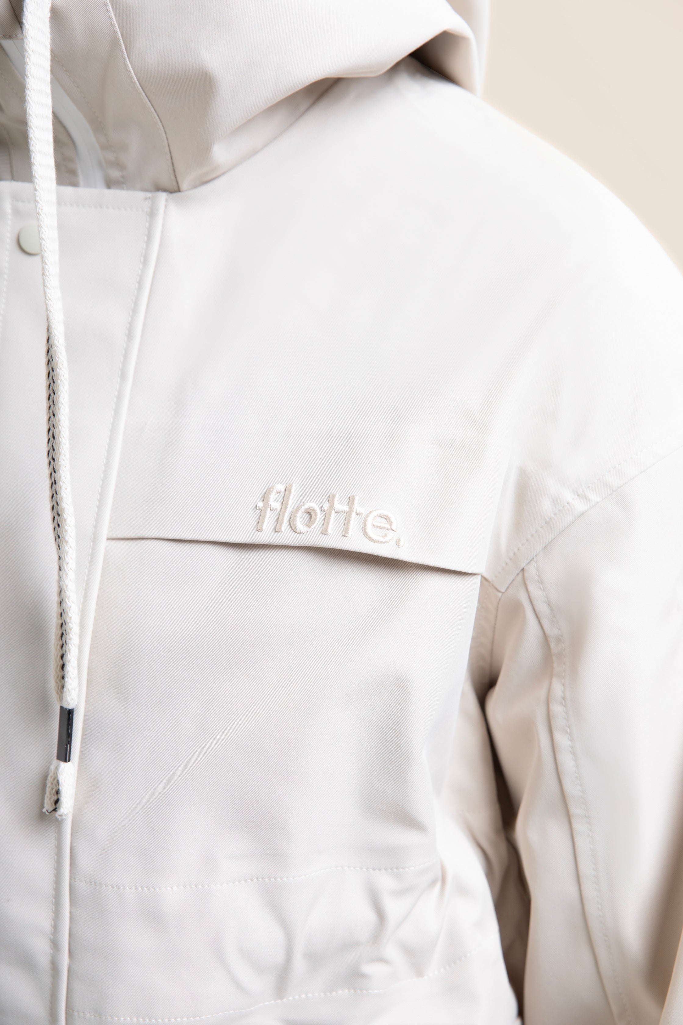 Close-up of a stylish beige Flotte waterproof parka coat featuring a hooded design and branded chest detail.