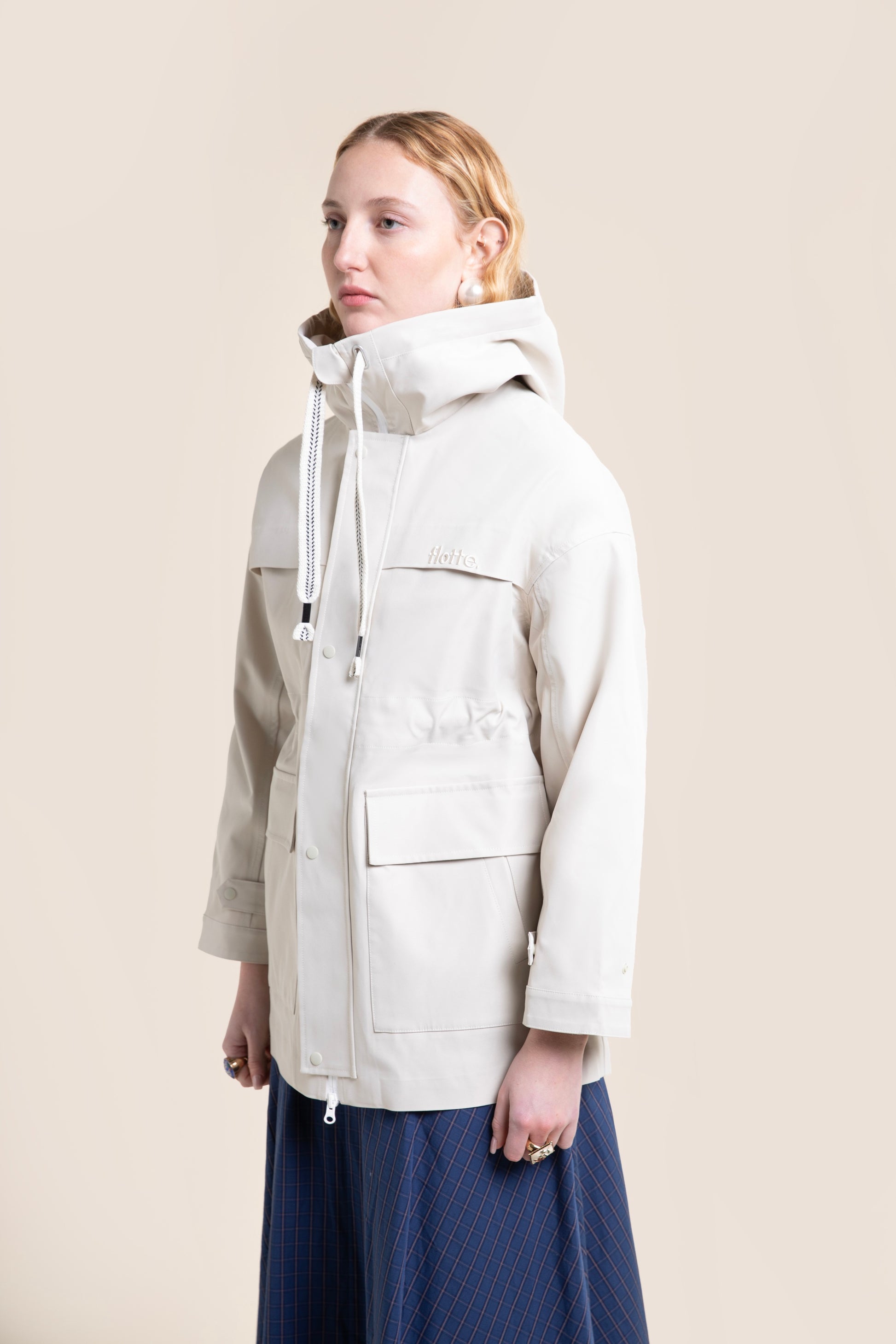 A person wearing a beige Flotte waterproof parka coat with a hood against a neutral background.