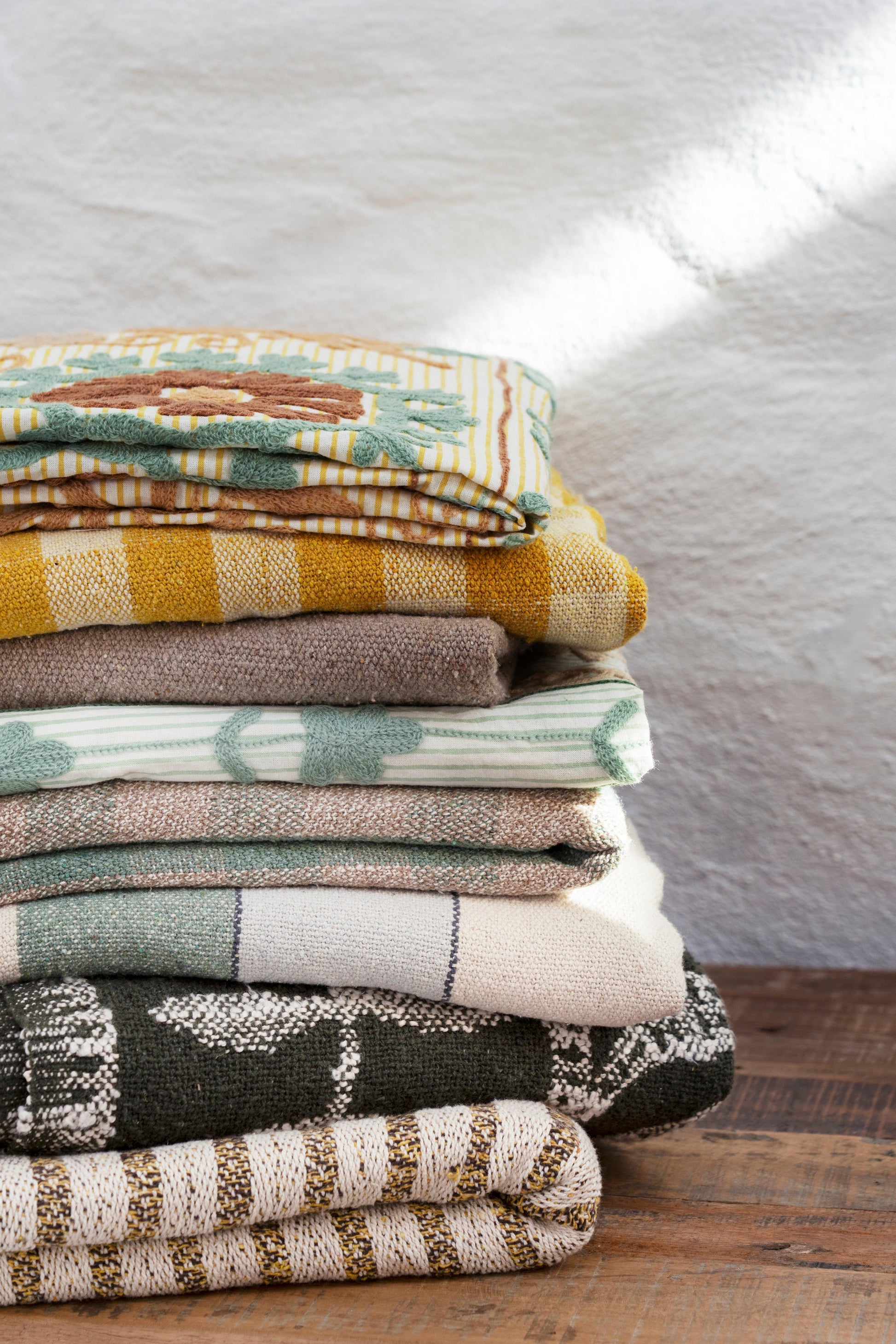 A neatly stacked pile of textured textile throws and blankets on a rustic wooden table against a white wall.