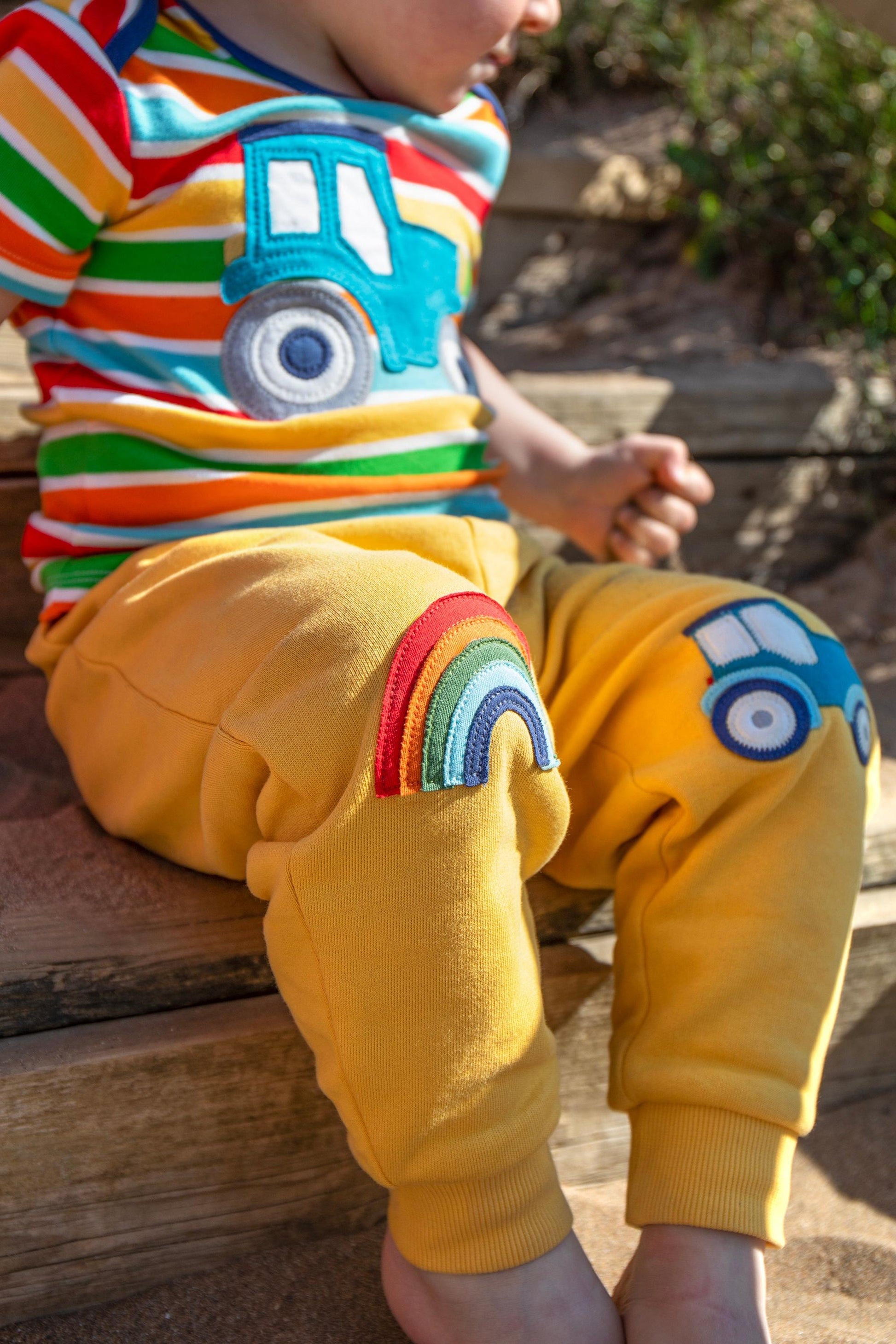 Child wearing a colorful shirt and yellow pants with car designs, sitting on wooden steps.