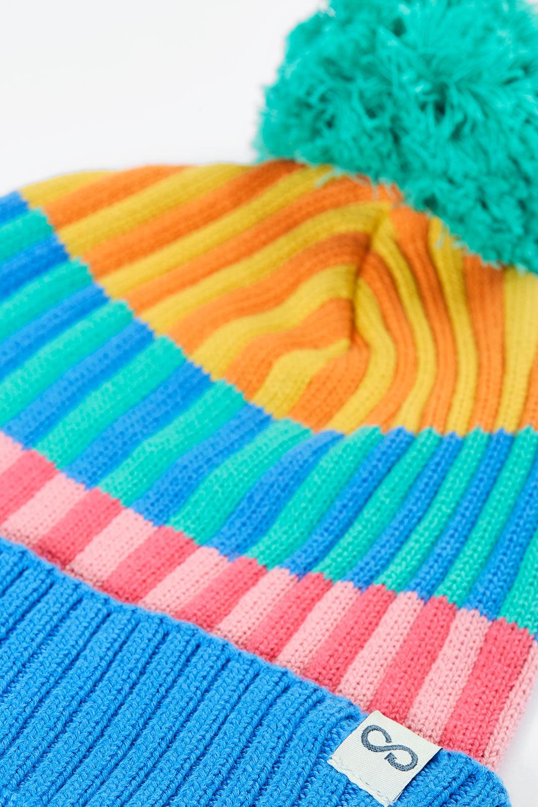 A close-up of a Frugi rainbow-striped knit beanie hat with a bright teal pom-pom