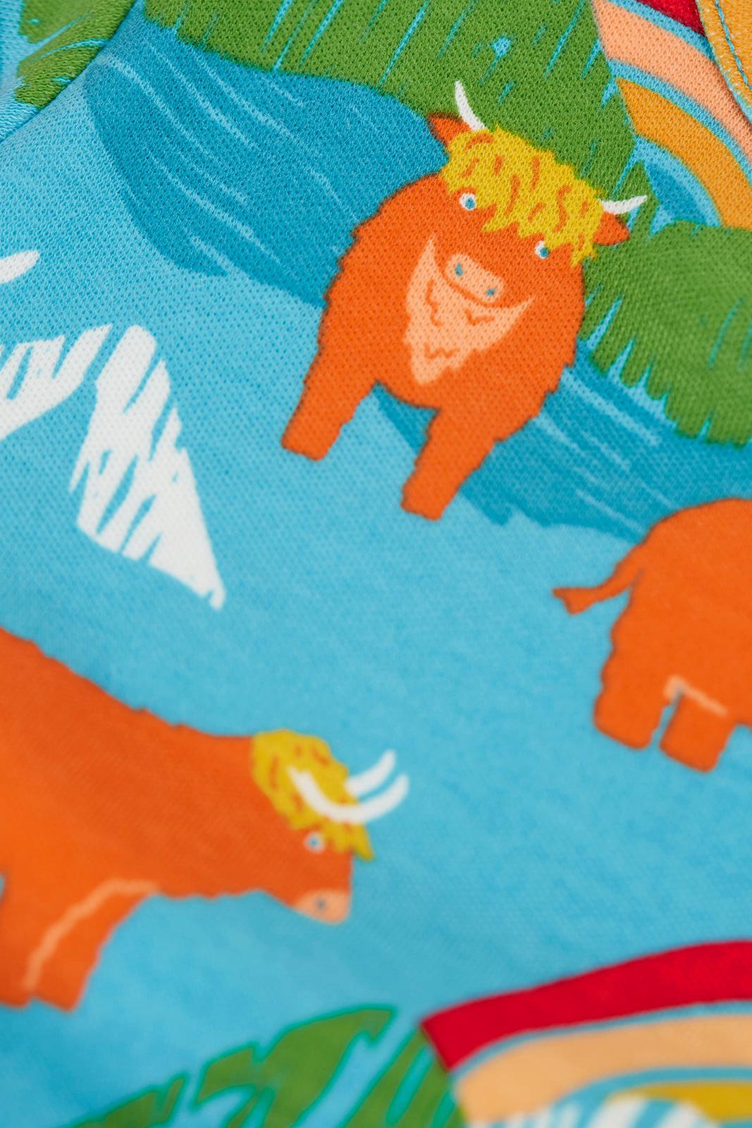 Close-up of a colorful printed fabric featuring cartoon orange Highland cows on a bright blue background with green leaves and white mountain shapes.