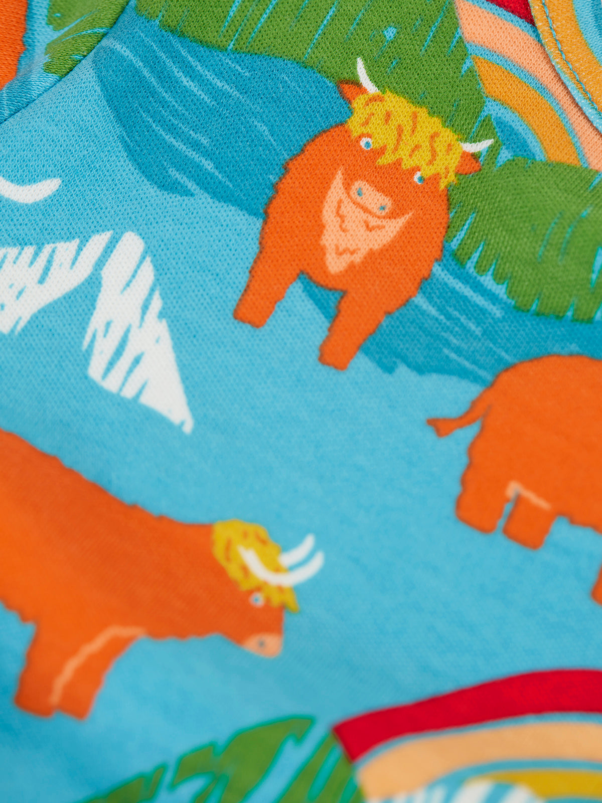 Close-up of a colorful printed fabric featuring cartoon orange Highland cows on a bright blue background with green leaves and white mountain shapes.
