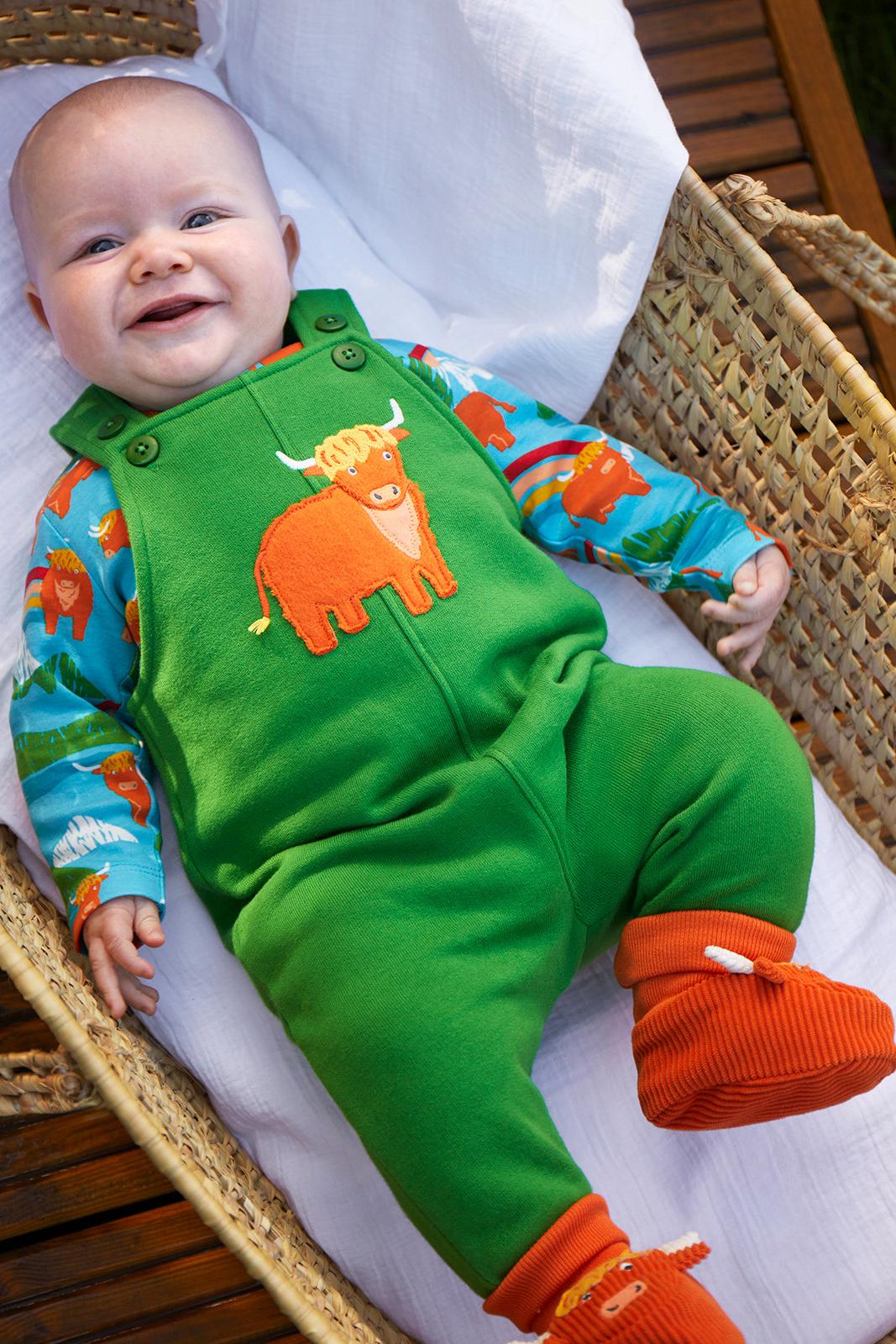 A smiling baby lying on a white cushion inside a woven wicker basket, wearing bright green overalls with an orange cow applique, a blue and orange animal-print long-sleeve top, and orange knit booties.
