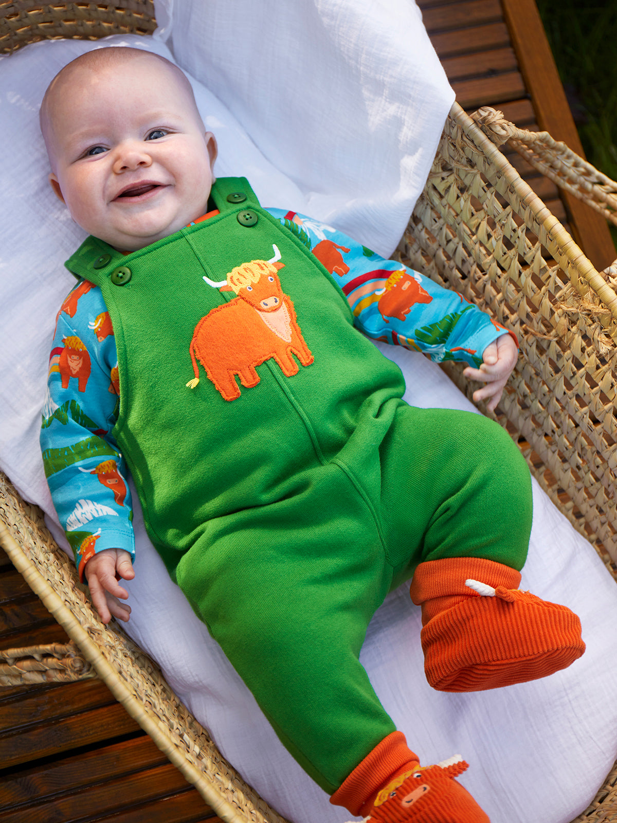 A smiling baby lying on a white cushion inside a woven wicker basket, wearing bright green overalls with an orange cow applique, a blue and orange animal-print long-sleeve top, and orange knit booties.