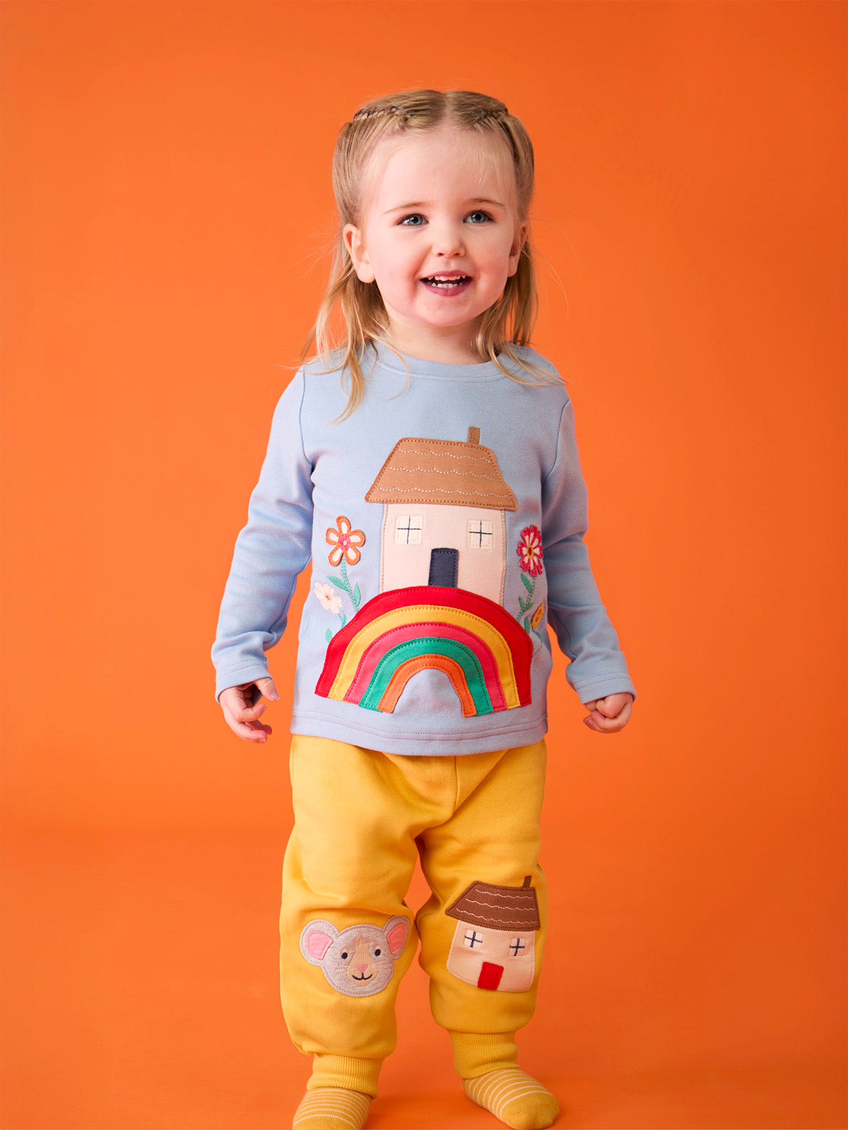 A happy toddler girl wearing a Frugi light blue organic cotton top with a rainbow and house applique design.
