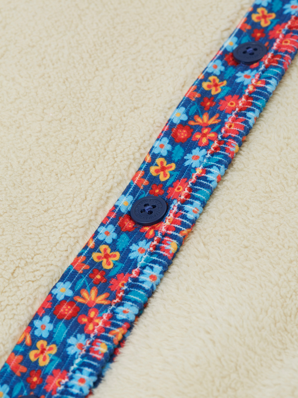 Close-up of a Frugi girls fleece jacket featuring a blue floral trim and dark blue buttons on cream-colored fabric.