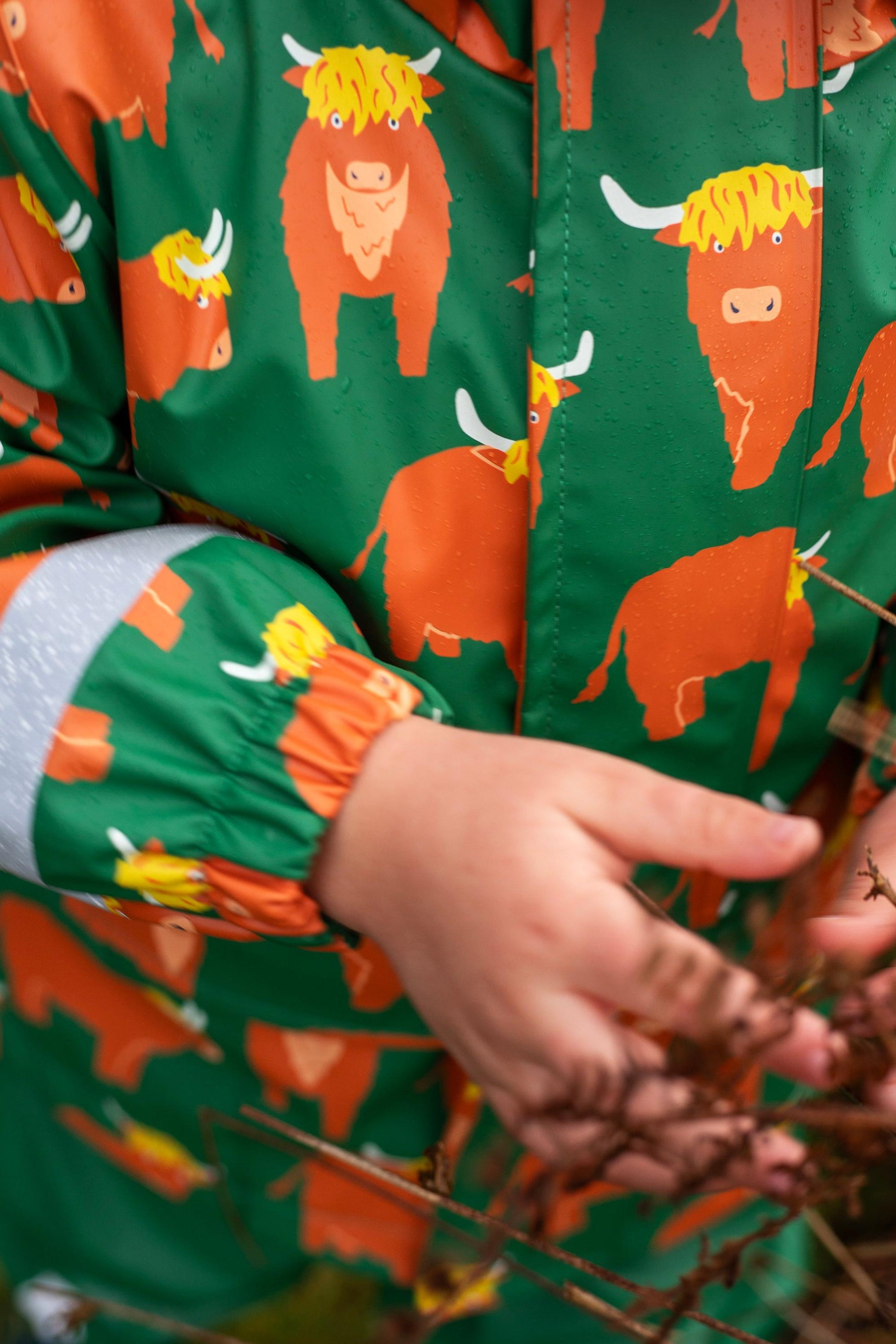 Child wearing a green Frugi waterproof jacket with orange highland cow print outdoors