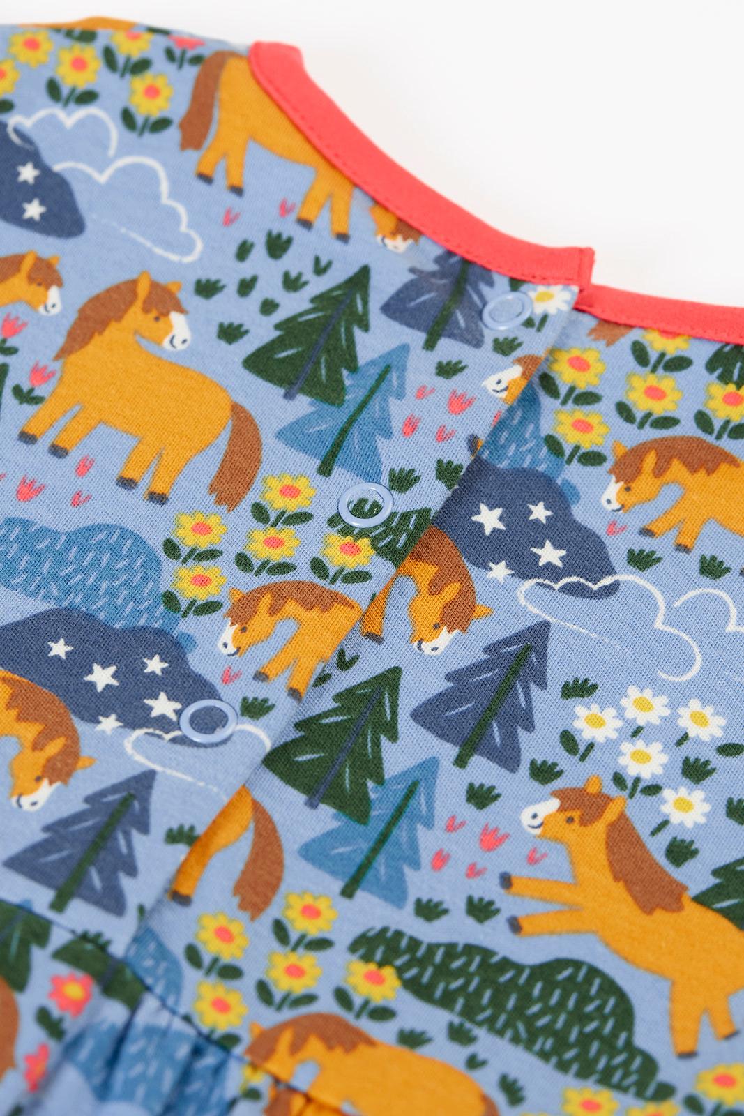 Close-up of a Frugi organic cotton baby dress featuring a colourful horse, flower, and forest pattern with snap buttons.