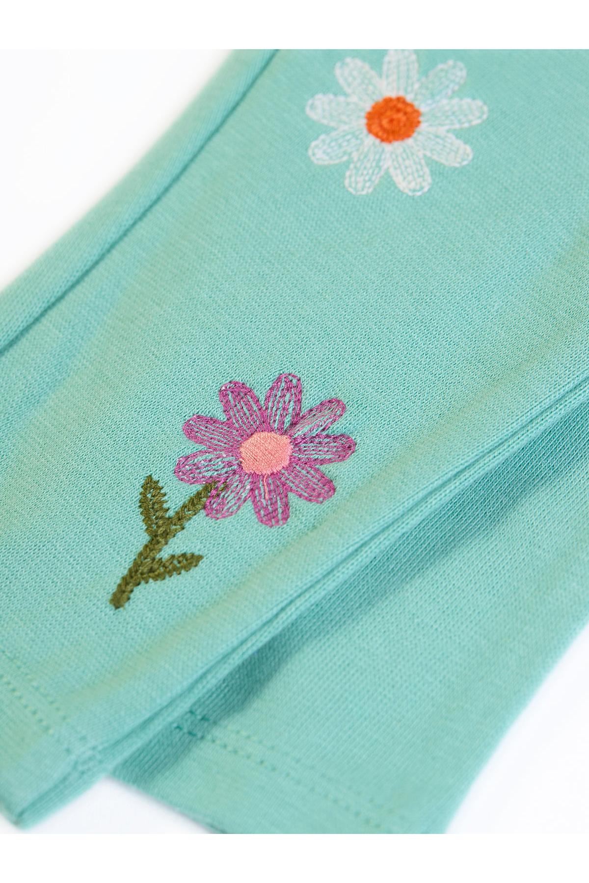 A close-up of a mint green fabric item, likely a pair of baby tights or a soft textile accessory, featuring embroidered daisy flowers in white, pink, and orange with green stems.