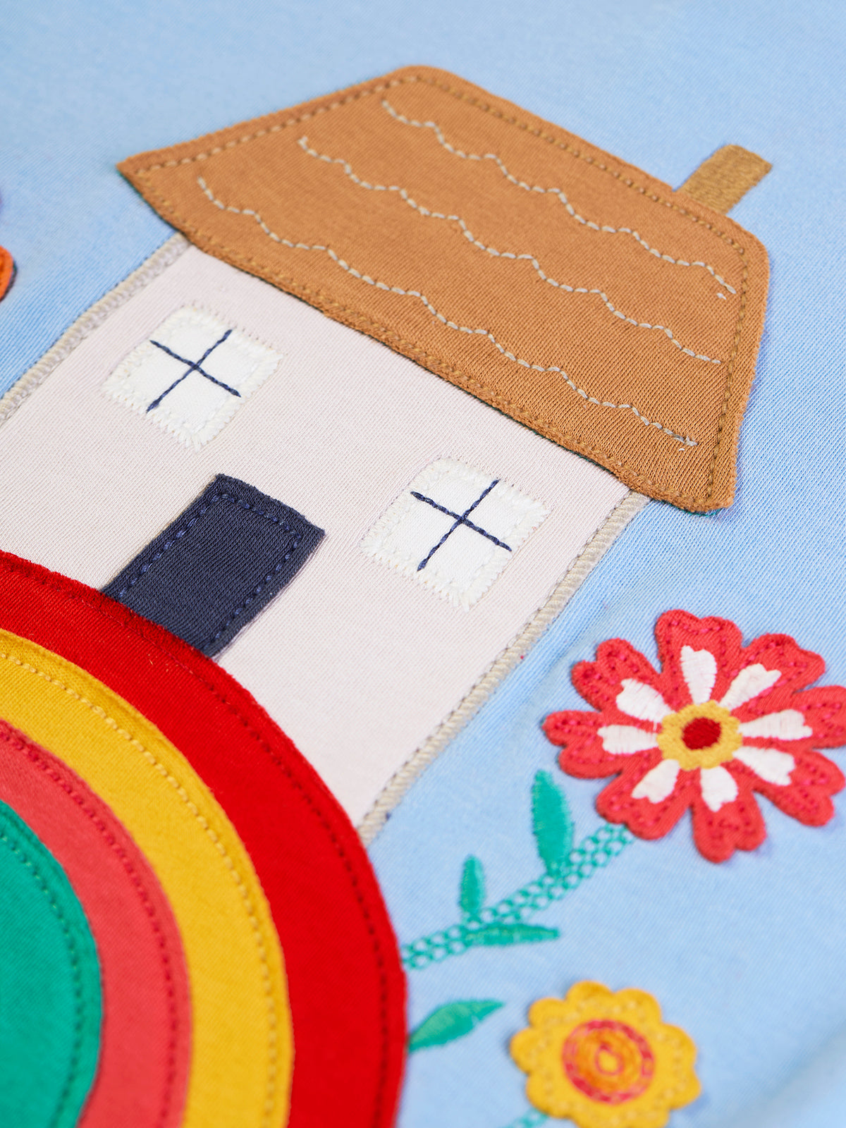 Close-up of a Frugi organic cotton girl's top with a colourful house and flower applique design on light blue fabric.