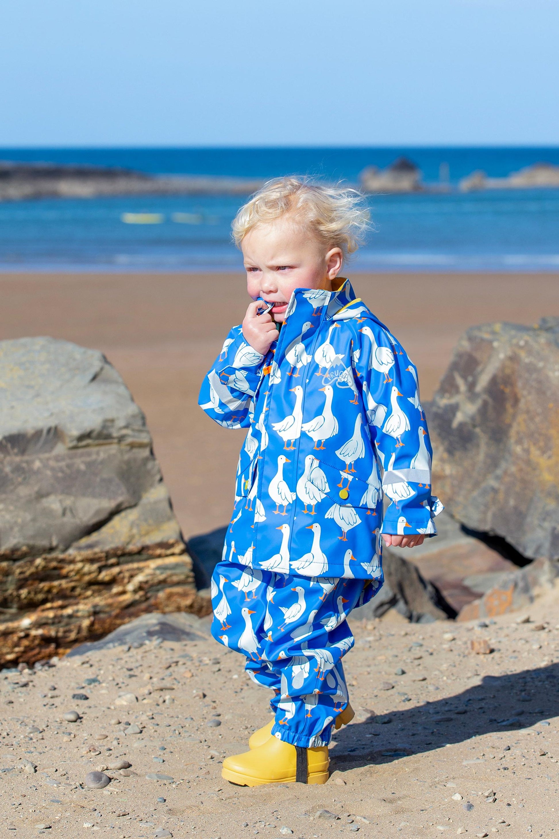Child wearing a blue Frugi raincoat with white goose pattern on a beach