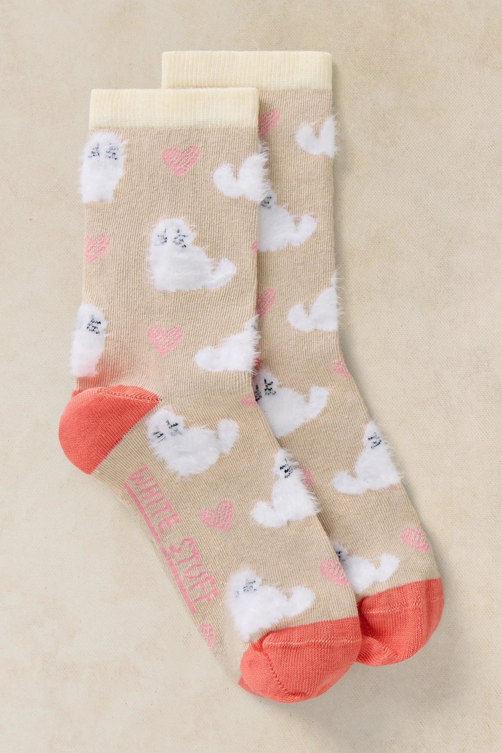 A pair of beige crew socks with a pattern of fluffy white cats, pink hearts, and coral orange toes and heels, displayed on a neutral beige background.