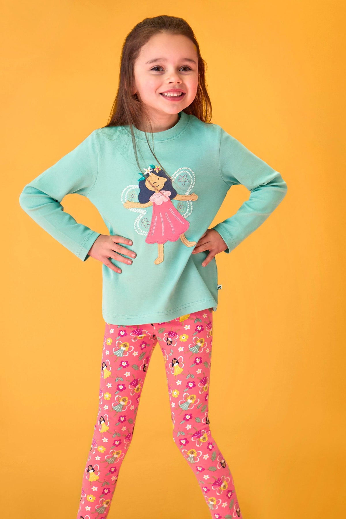 A smiling young girl posing in a teal long-sleeve top and pink fairy-print organic cotton girls leggings.