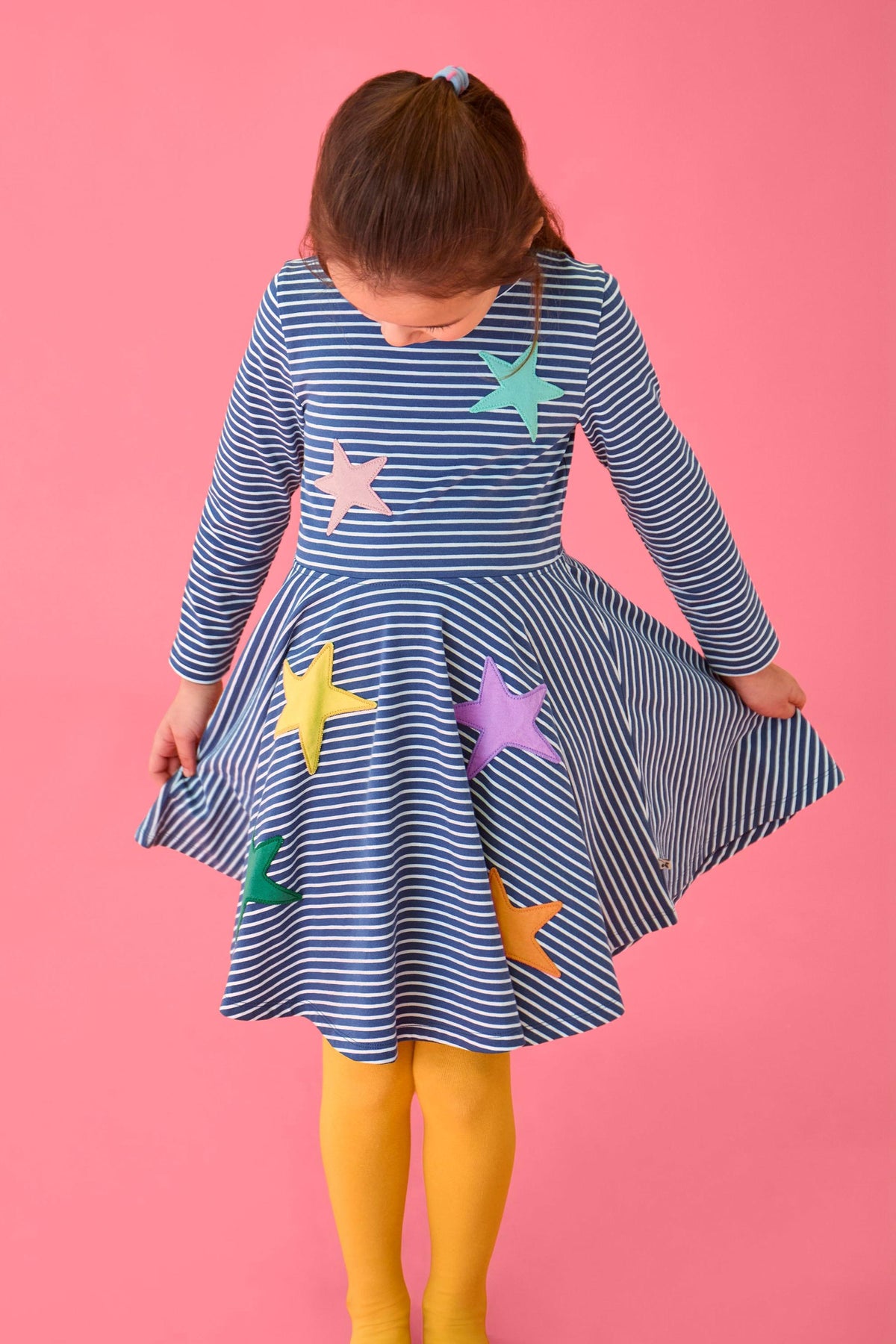 A young girl wearing a blue and white striped organic cotton Frugi spinning dress with colorful star appliques.