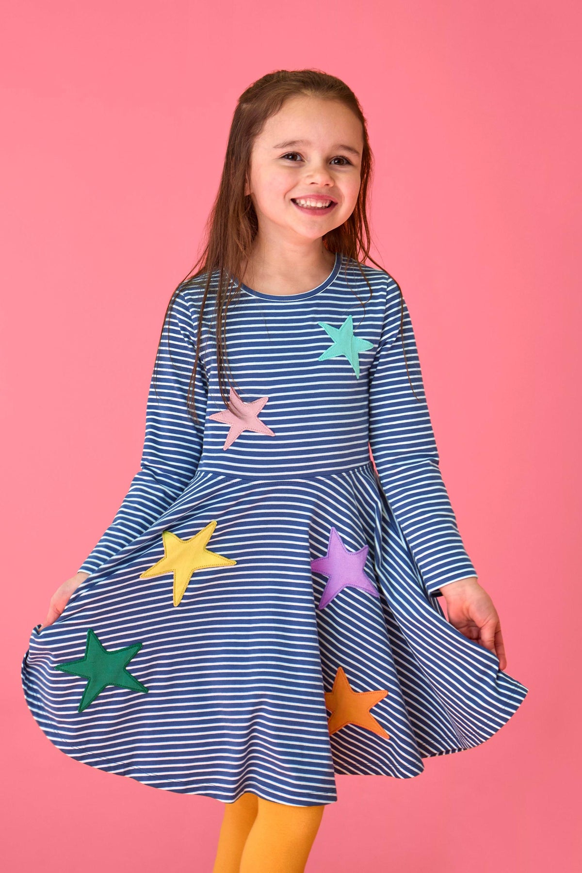 Young girl smiling while wearing a navy stripe Frugi spinning dress with colorful stars against a pink background.
