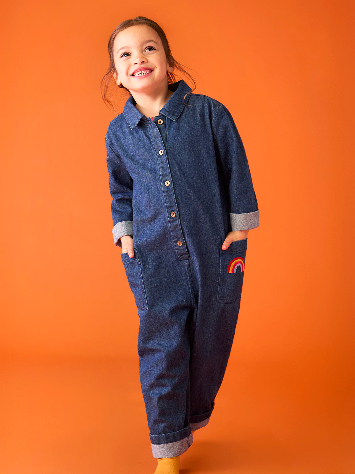 Smiling girl wearing a Frugi organic denim jumpsuit with a colourful rainbow detail on the pocket.