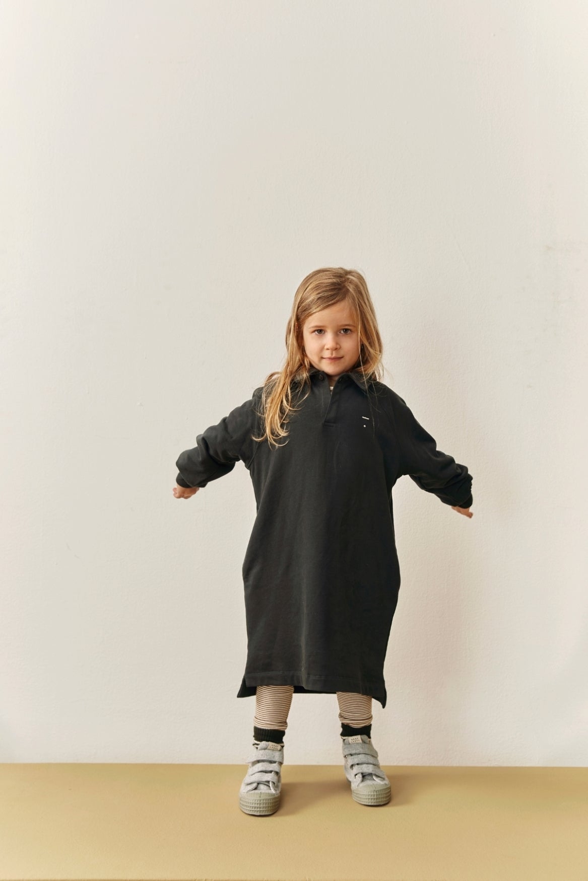 A young child wearing a long-sleeve black polo shirt dress, standing against a plain off-white wall and beige floor.
