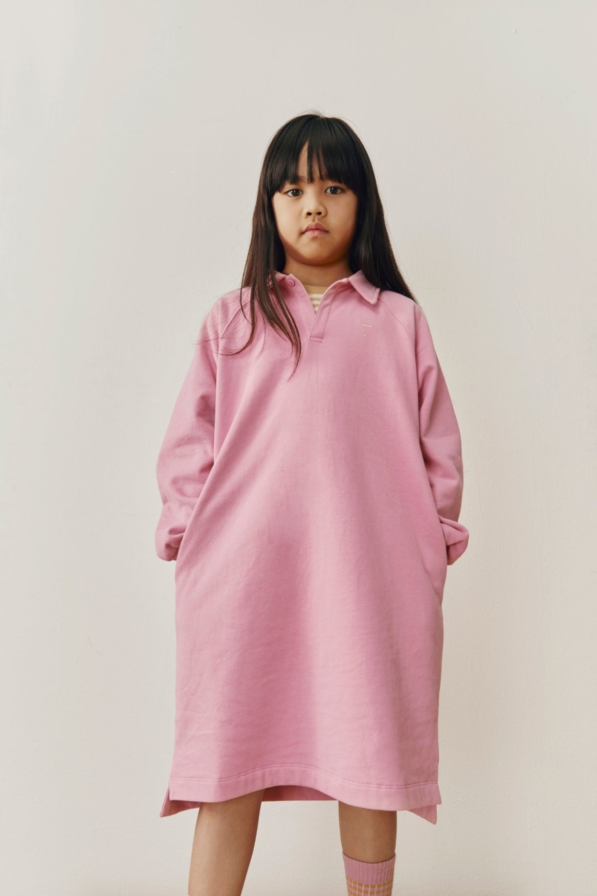 A young girl with long dark hair wearing an oversized pink long-sleeve polo shirt dress, standing against a plain light beige studio background.