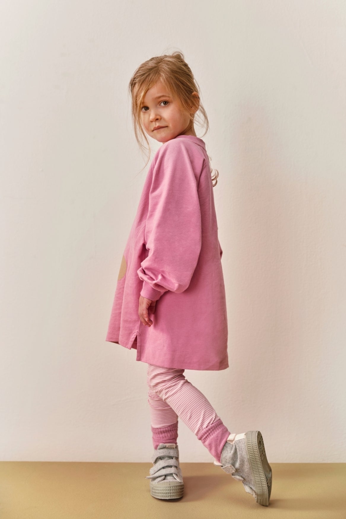 Young girl wearing a pink Gray Label organic cotton dress and striped leggings standing against a neutral background