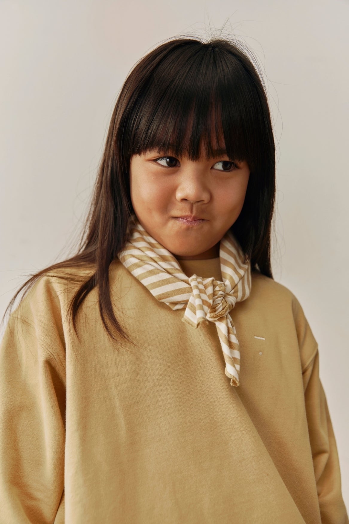 A young girl with dark hair and bangs wearing a beige long-sleeve top and a cream and beige striped neck scarf tied in a knot, photographed against a plain light beige background.