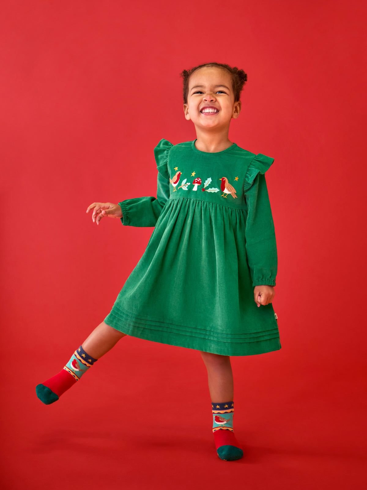 A happy child wearing a green Frugi organic cotton corduroy Christmas dress with robin embroidery.