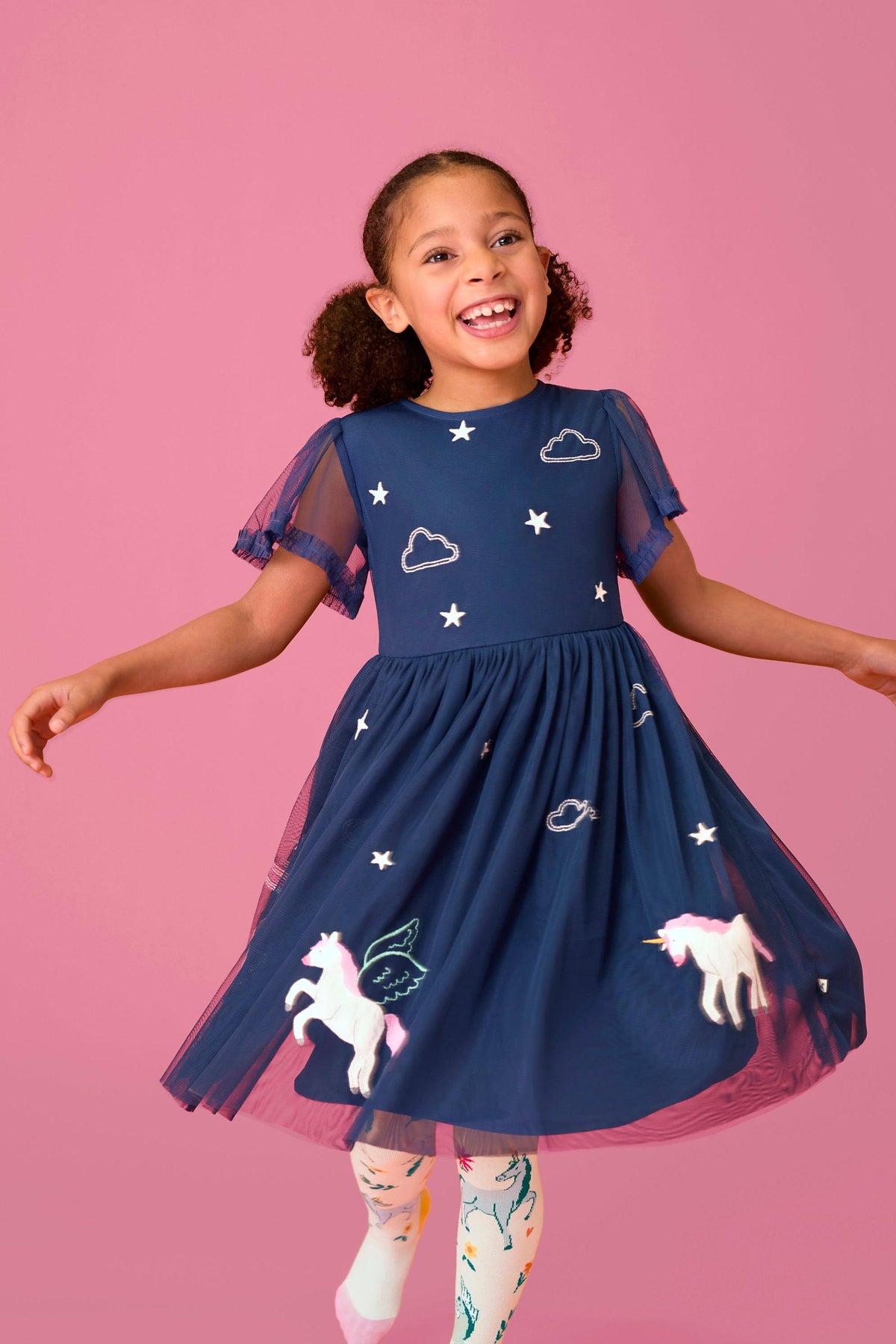 A joyful girl spinning in a navy Frugi unicorn party dress made of recycled polyester against a pink background.